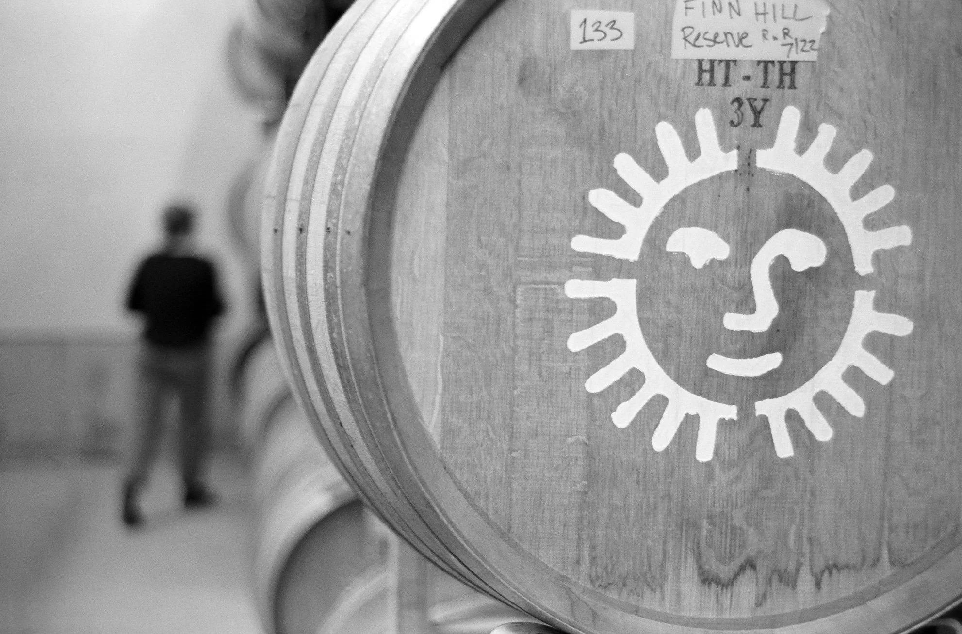 Close-up of a wooden wine barrel with our Fossil & Fawn. In the background, a person stands near more barrels, out of focus.