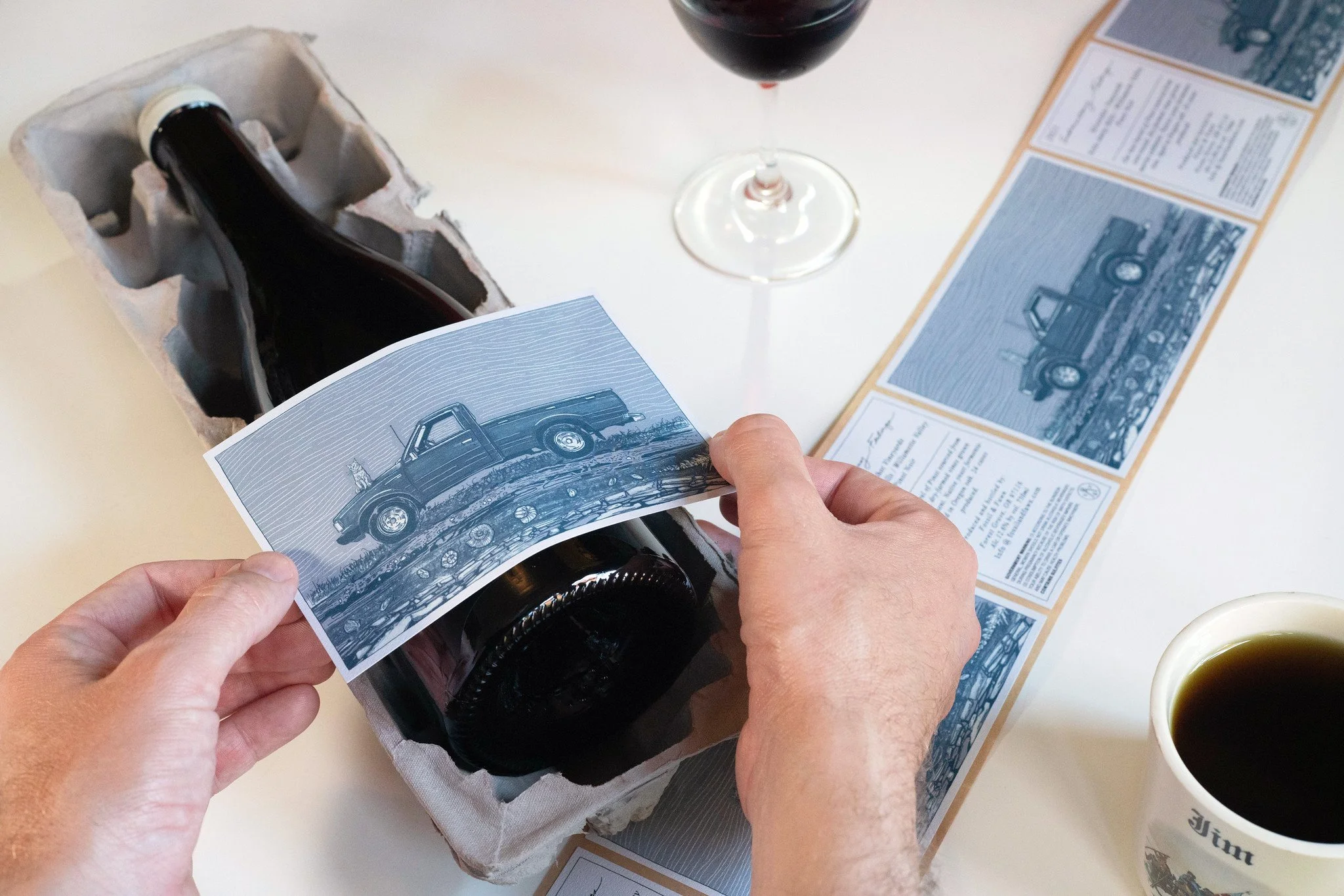 Person holding a black and white photo of a vintage truck over a bottle of our "Sedimentary Feelings" Pinot noir in a cardboard carrier. A glass of red wine, a long paper menu, and a cup of coffee are on a white table.