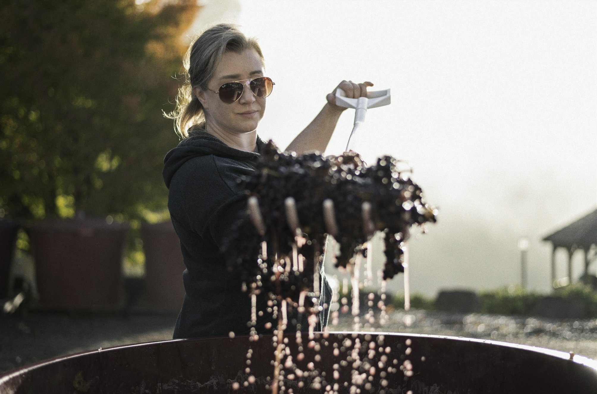 Woman wearing sunglasses and a black hoodie crushing grapes in a large container with a tool outdoors during sunrise.