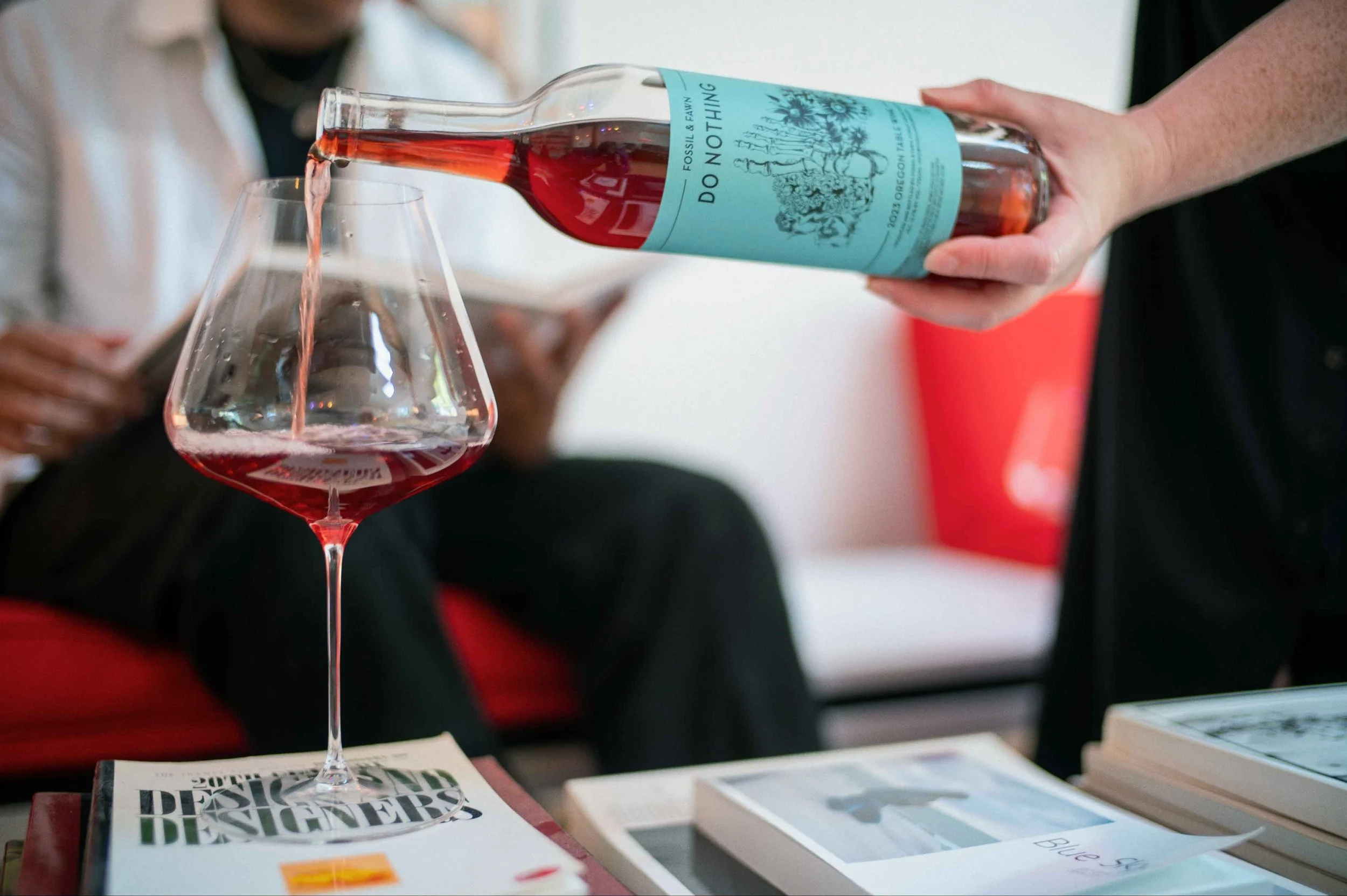 Person pouring our Do Nothing wine into a glass on a table with magazines and books, while another person sits in the background.