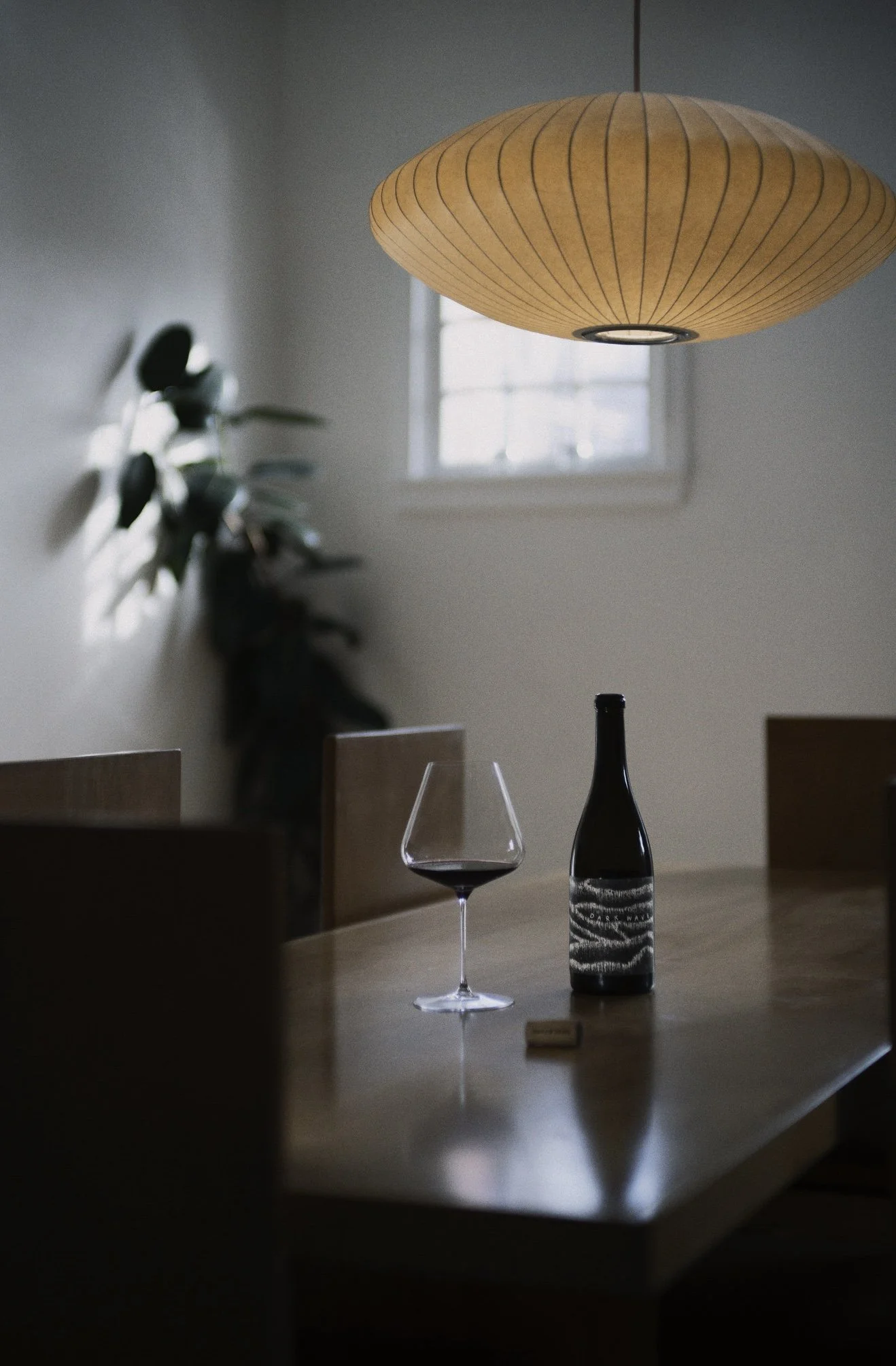 A wooden dining table with a glass of our "Dark Wave" red wine and a wine bottle, illuminated by a hanging orange paper lantern, in a room with a window and a plant in the background.