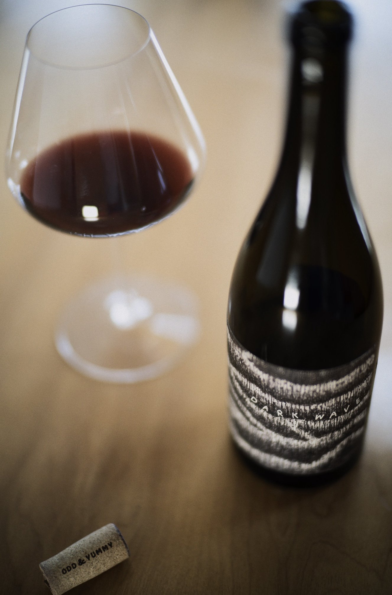 A wine glass filled with our "Dark Wave" red wine, a dark-colored wine bottle with a black and white label, and a small cork with the words 'Odd & Yummy!' on a wooden surface.