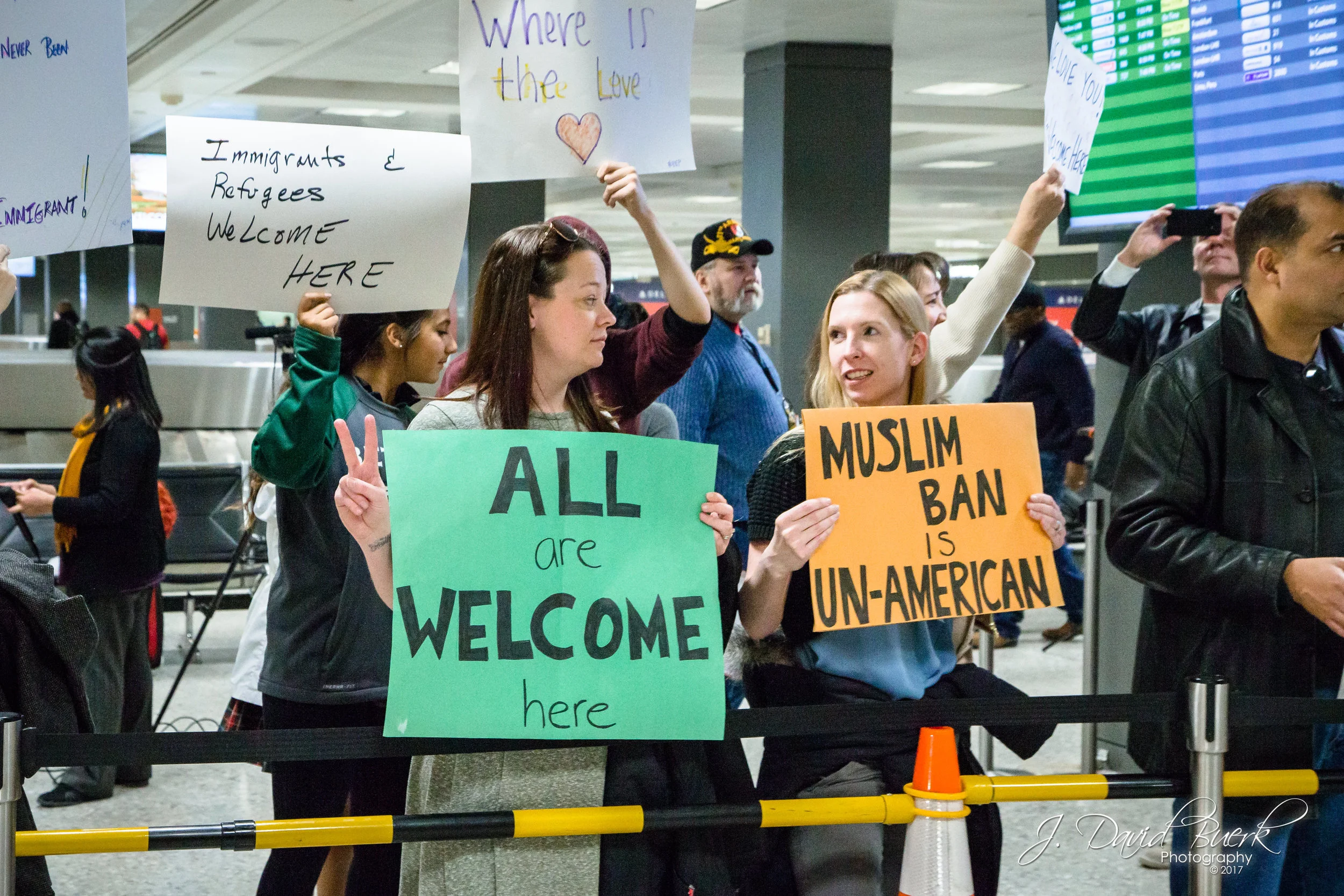 A woman shows a peace sign while holding a sign; "ALL are WELCOME here."
