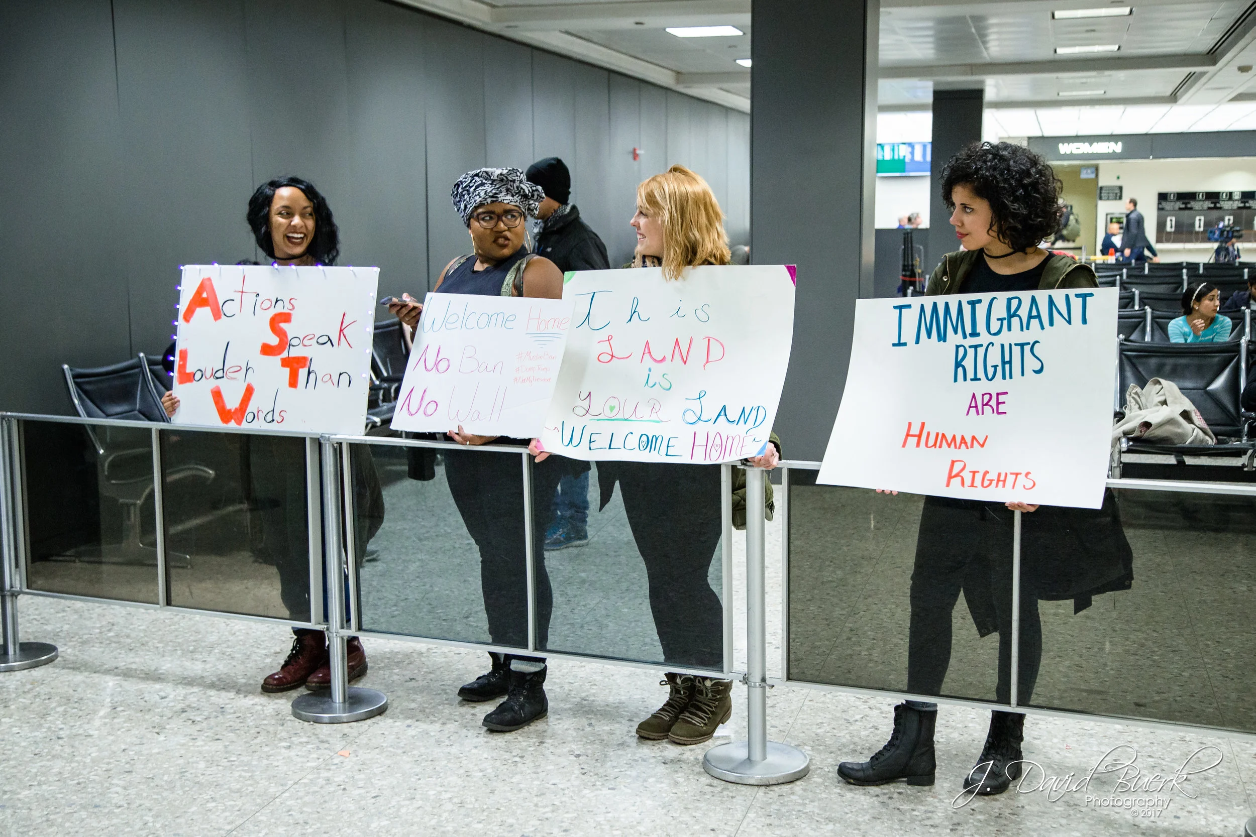 Four women hold signs next to the International Arrival Building's exit; one woman's sign had flashing lights, reading, "Actions Speak Louder Than Words."