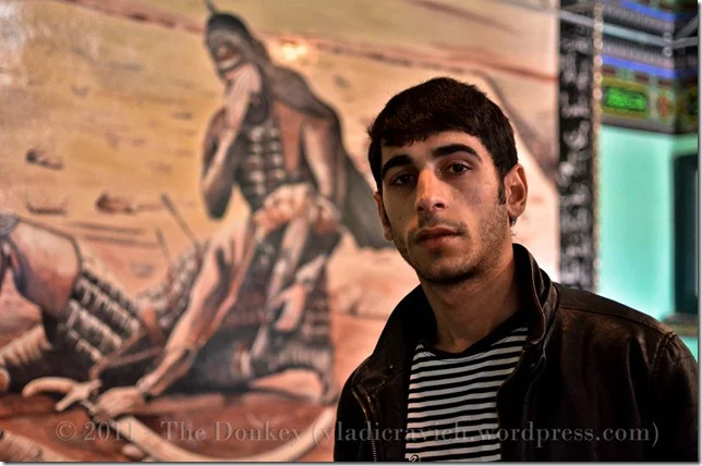 Suleman Kindir in his family's pir, in front of a mural added in the last few years depicting the death of Imam Hussein, a pivotal moment in Shii Islam.