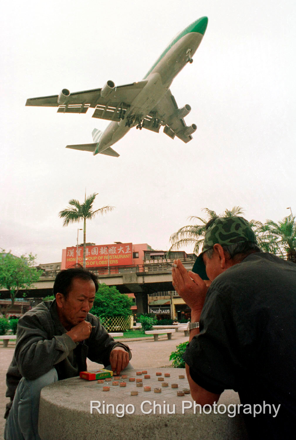  Two seniors play Chinese chess while an airplane is flying over in Kowloon City, Hong Kong.(1994) 