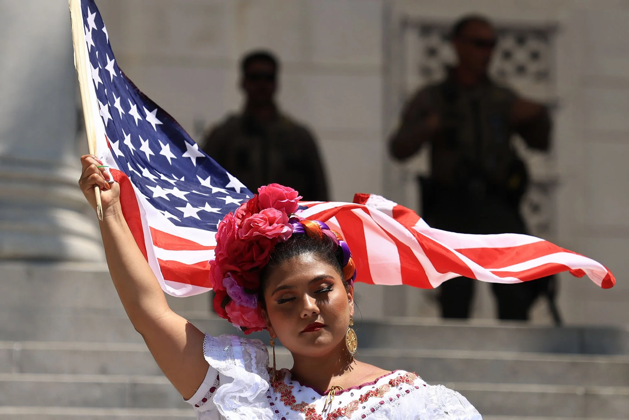  A Mexican dancer waves a U.S. flag as armed officers stand on the Los Angeles City Hall steps during a protest against federal immigration sweeps, in downtown Los Angeles, California. 