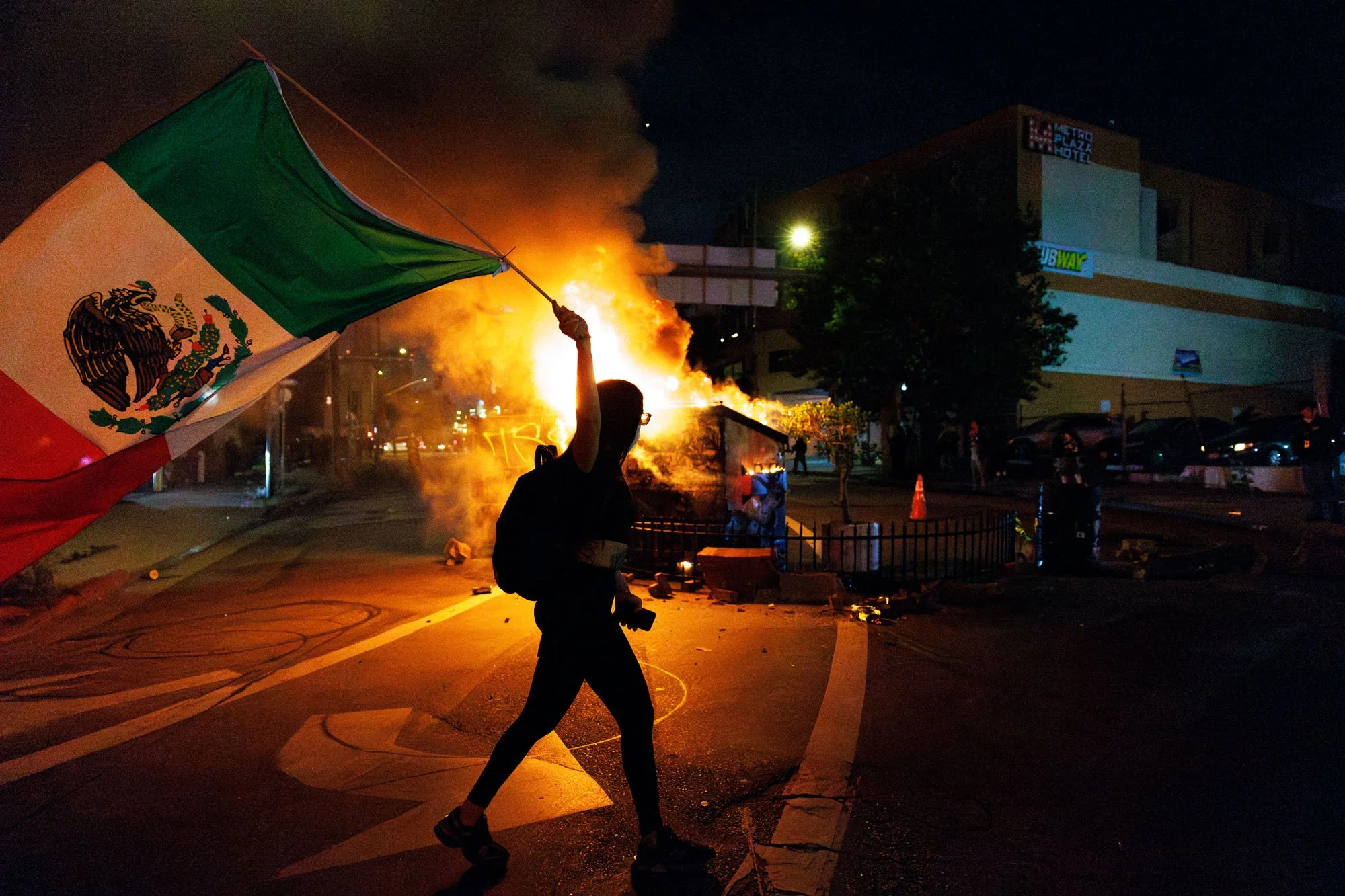  LOS ANGELES, CA - A demonstrator waves a Mexican flag in front  of a dumpster fire after another night of unrest during a protest against immigration raids on June 8, 2025 in Los Angeles, California. 
