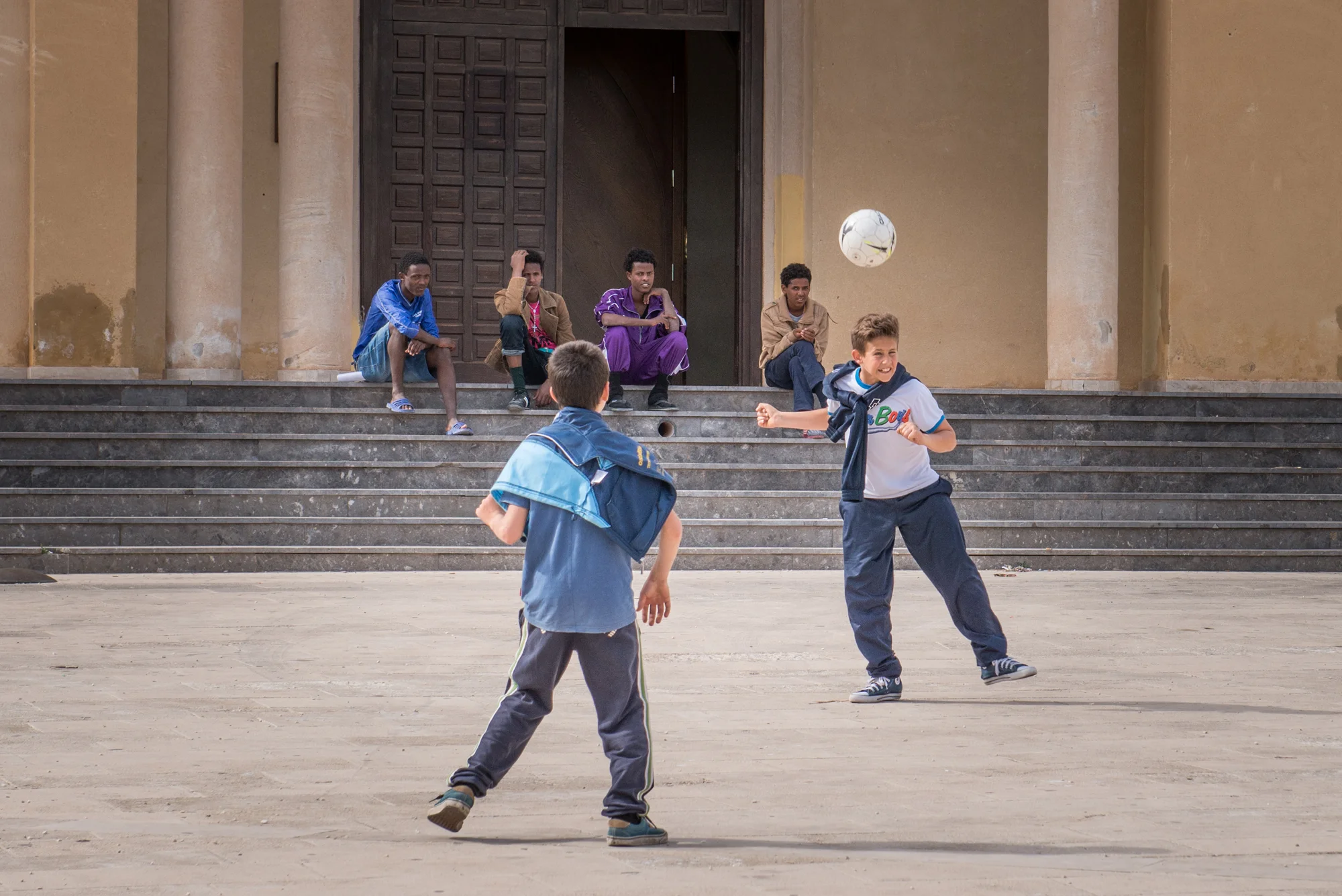 Young refugees sit on the church steps and watch a group of Italian kids playing football.