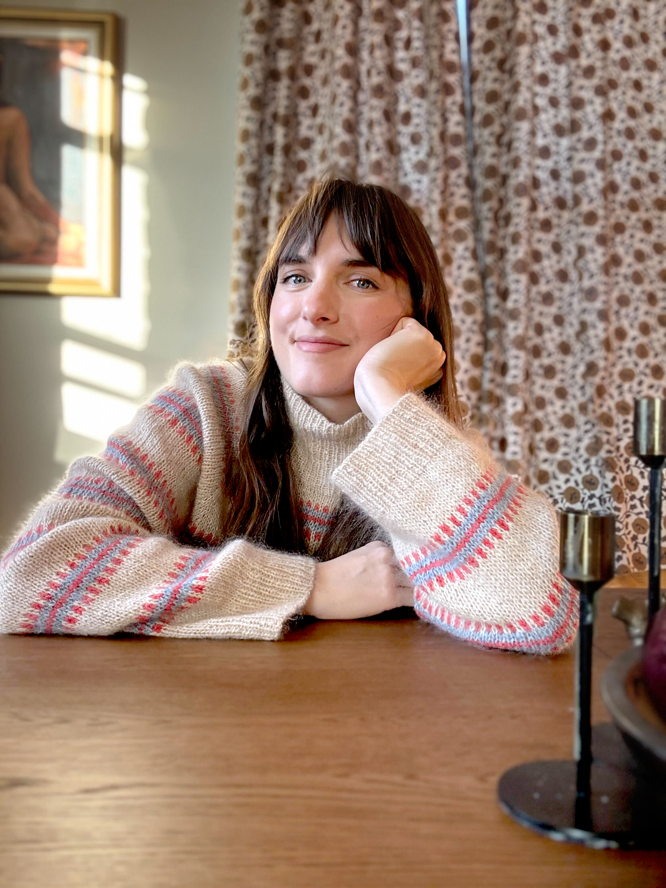 A young woman with long brown hair and blue eyes, smiling with her chin resting on her hand, wearing a beige sweater with colorful stripes, sitting at a wooden table in a cozy room with patterned curtains and a painting on the wall.