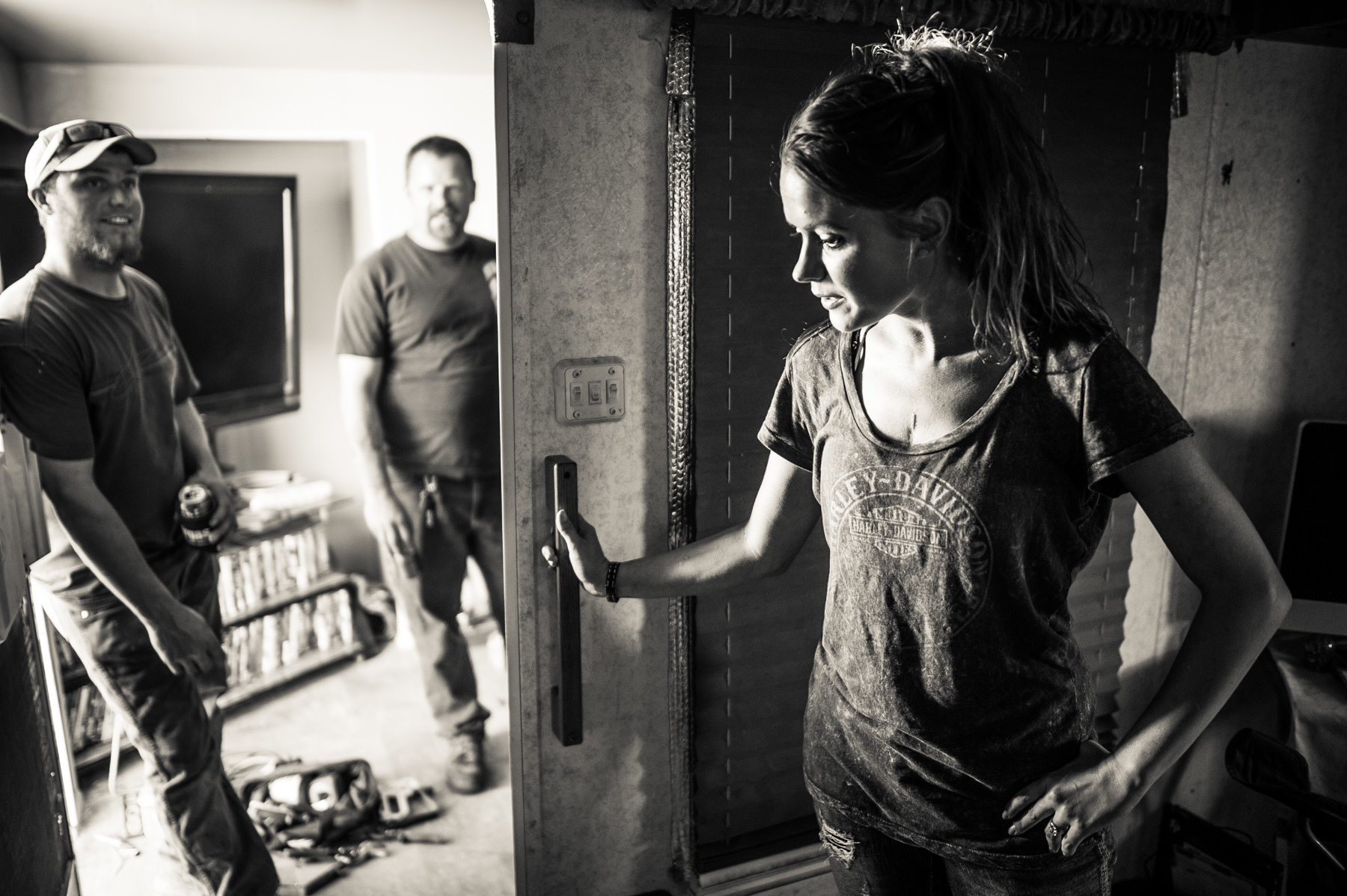  A woman, wife to an oil industry worker, stands in her travel trailer home while her husband and his friend talk and drink beer in the adjacent makeshift den. 
