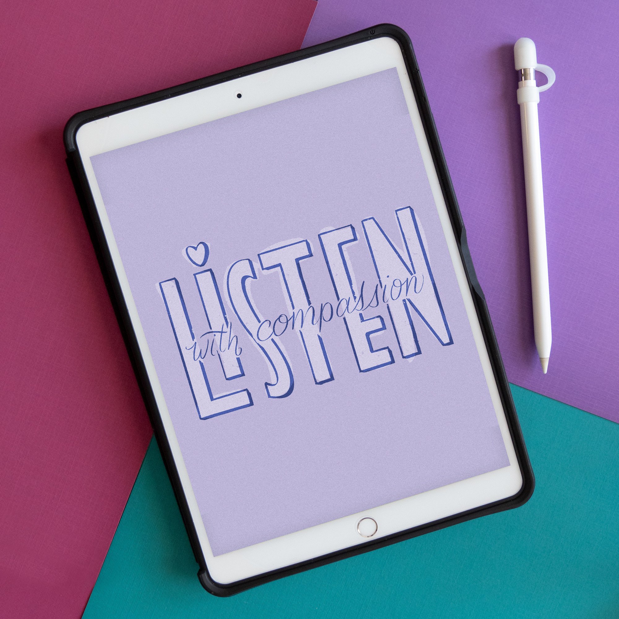 Tablet screen displaying a digital drawing with the handlettering "Listen with compassion," on a purple background, placed on a background of pink, purple, and teal papers with a white Apple Pencil beside it.
