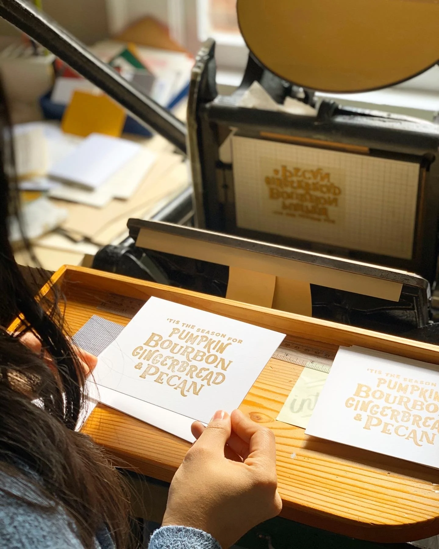 Person holding a printed card that reads 'Tis the season for pumpkin bourbon gingerbread & pecan' with a letterpress machine in the background.
