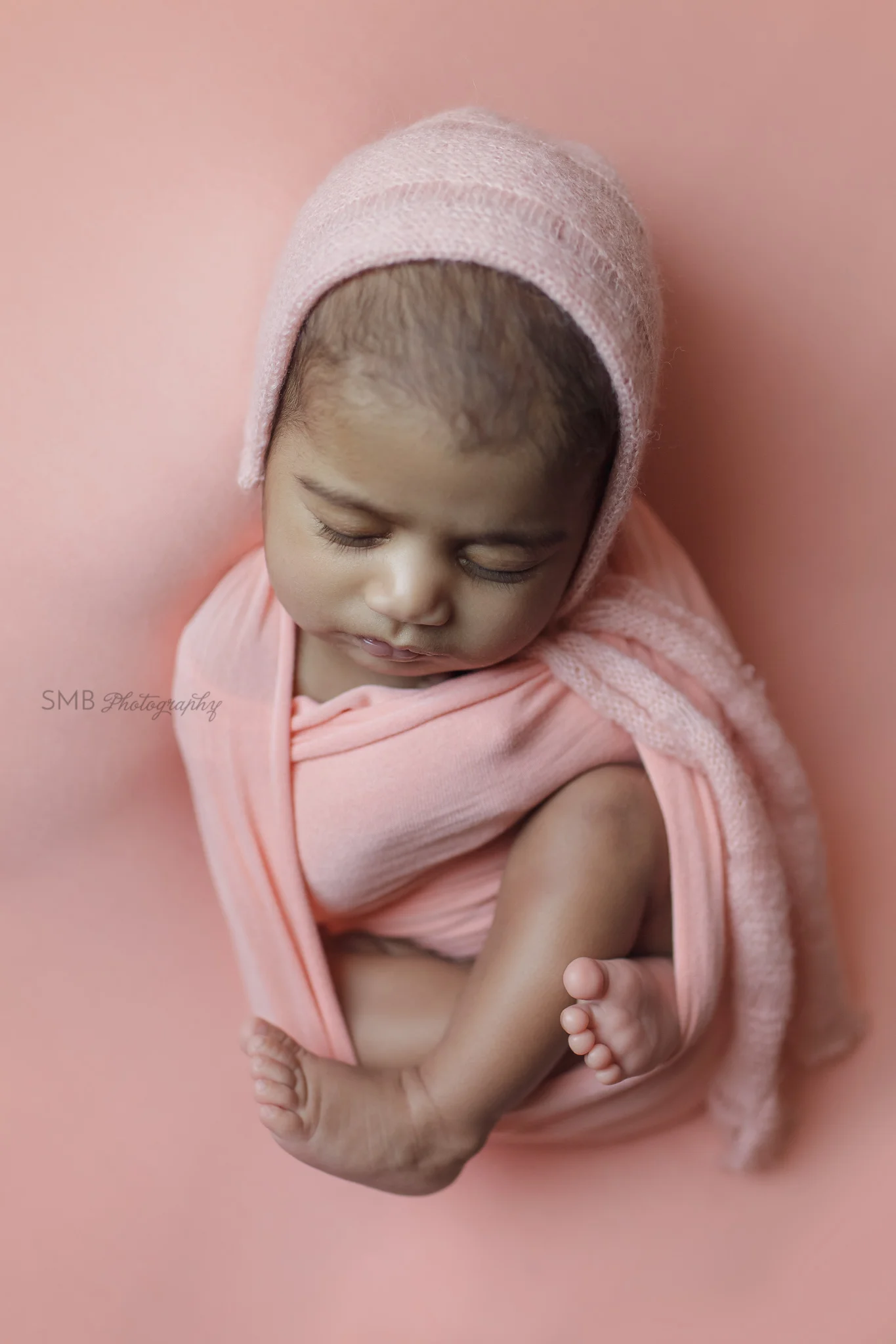 Newborn girl wrapped on peach backdrop, wearing peach bonnet