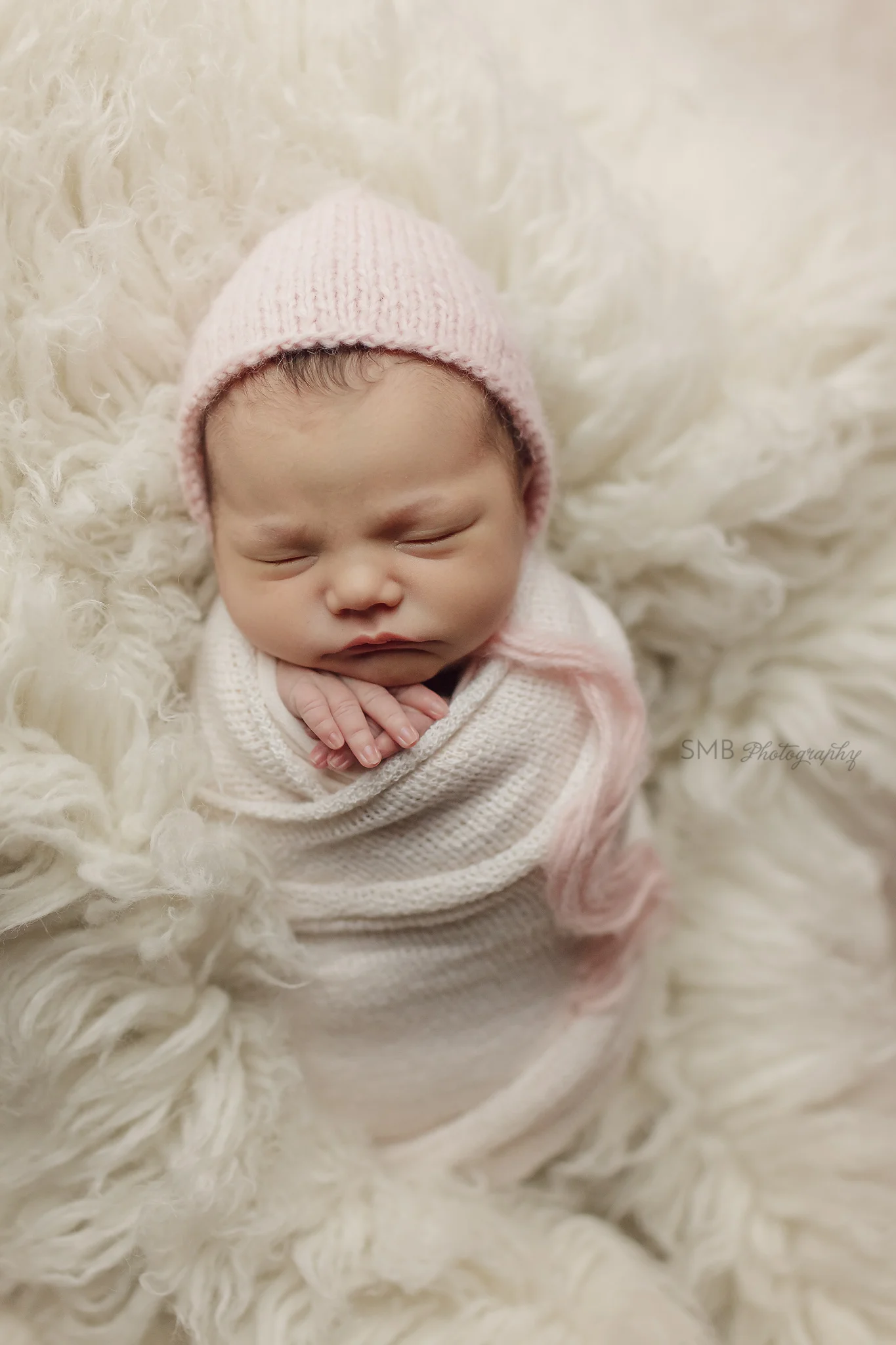 Newborn girl wearing pink bonnet swaddled on flokati