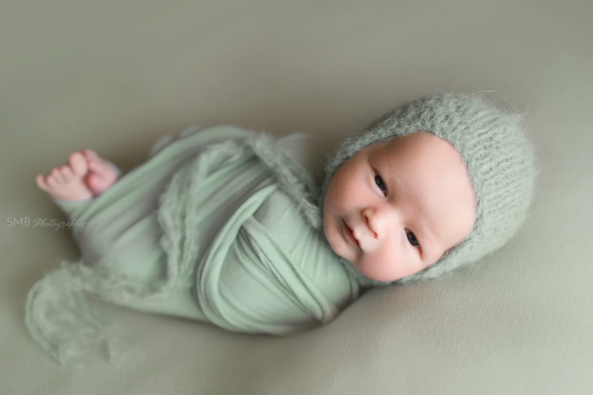 Newborn wrapped on sage green fabric wearing fuzzy bonnet