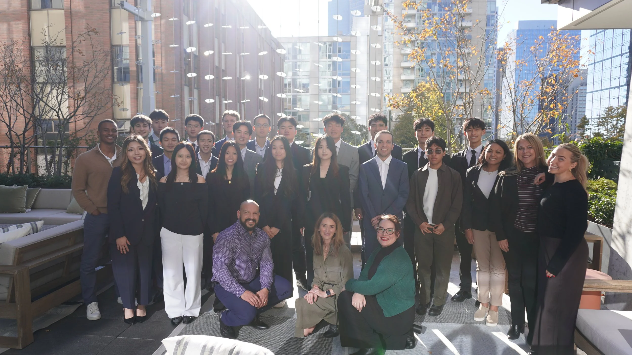 Group of diverse people, including men and women, posing for a photo outdoors on a rooftop terrace with modern buildings and sunny weather in the background.