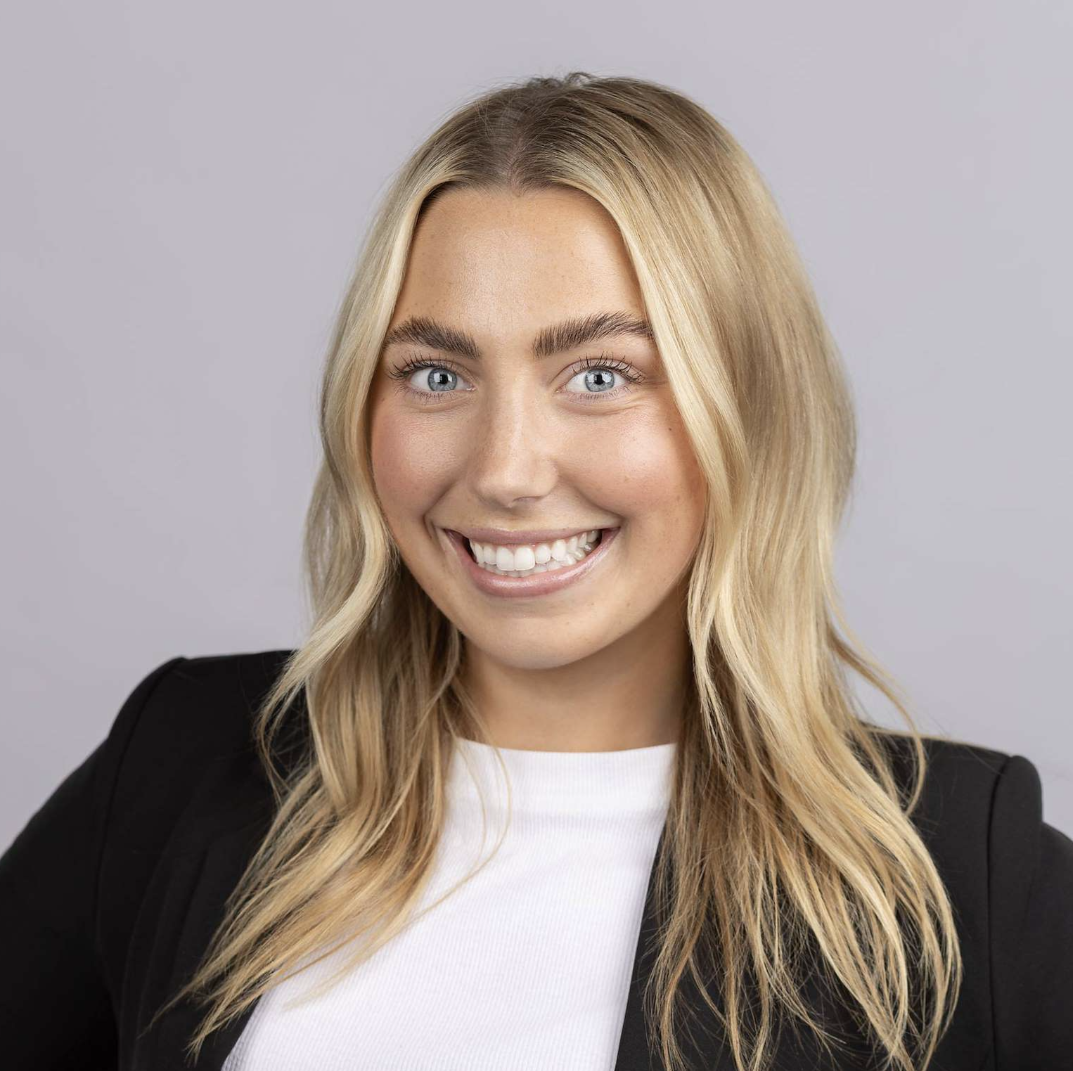 A smiling woman with blonde hair and blue eyes, wearing a black blazer and white top, against a plain gray background.