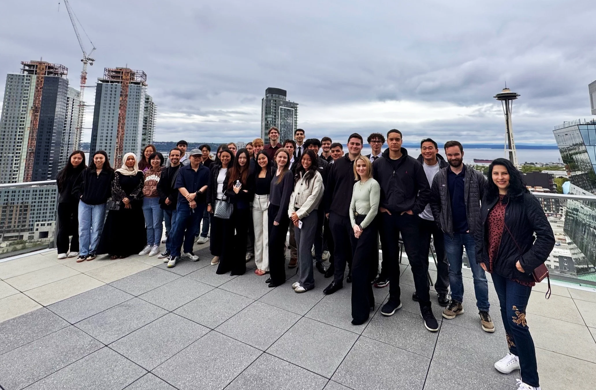 A diverse group of people, including men and women of various ethnicities, standing together on a rooftop with a city skyline, including the Space Needle and high-rise buildings, in the background during daytime with cloudy skies.