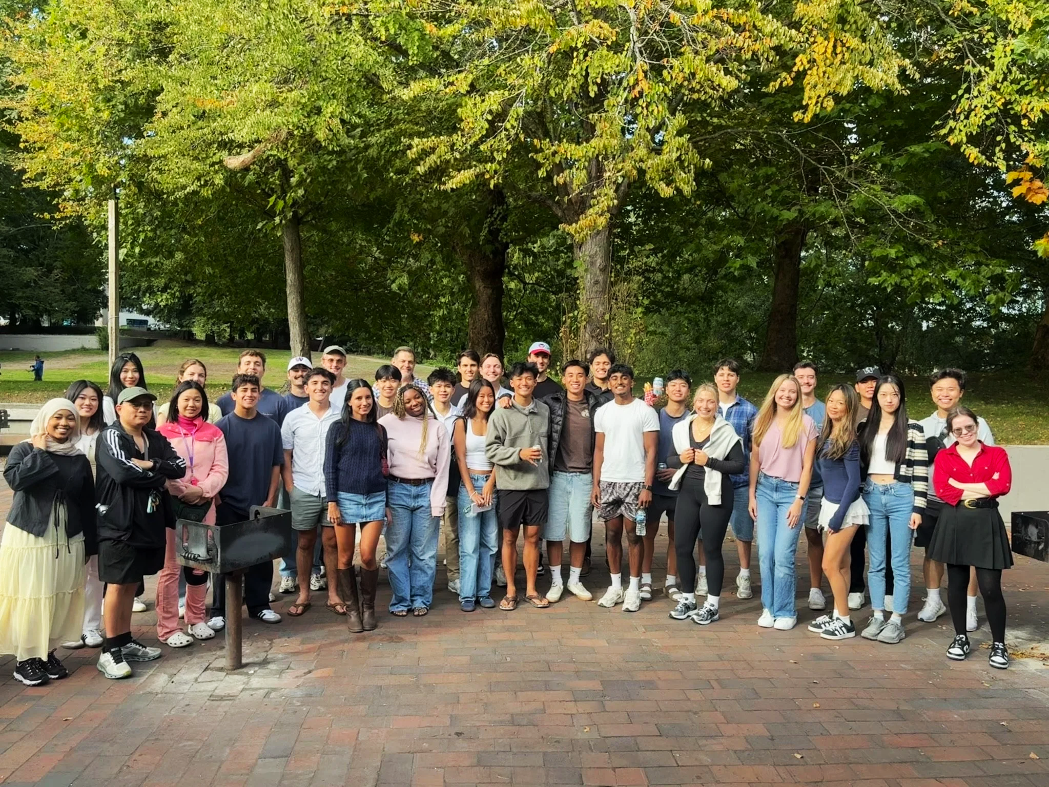 A large group of diverse young people gathered outdoors in a park, posing for a group photo under a large leafy tree with some people holding drinks. The group includes males and females of various ethnicities, dressed casually, smiling at the camera