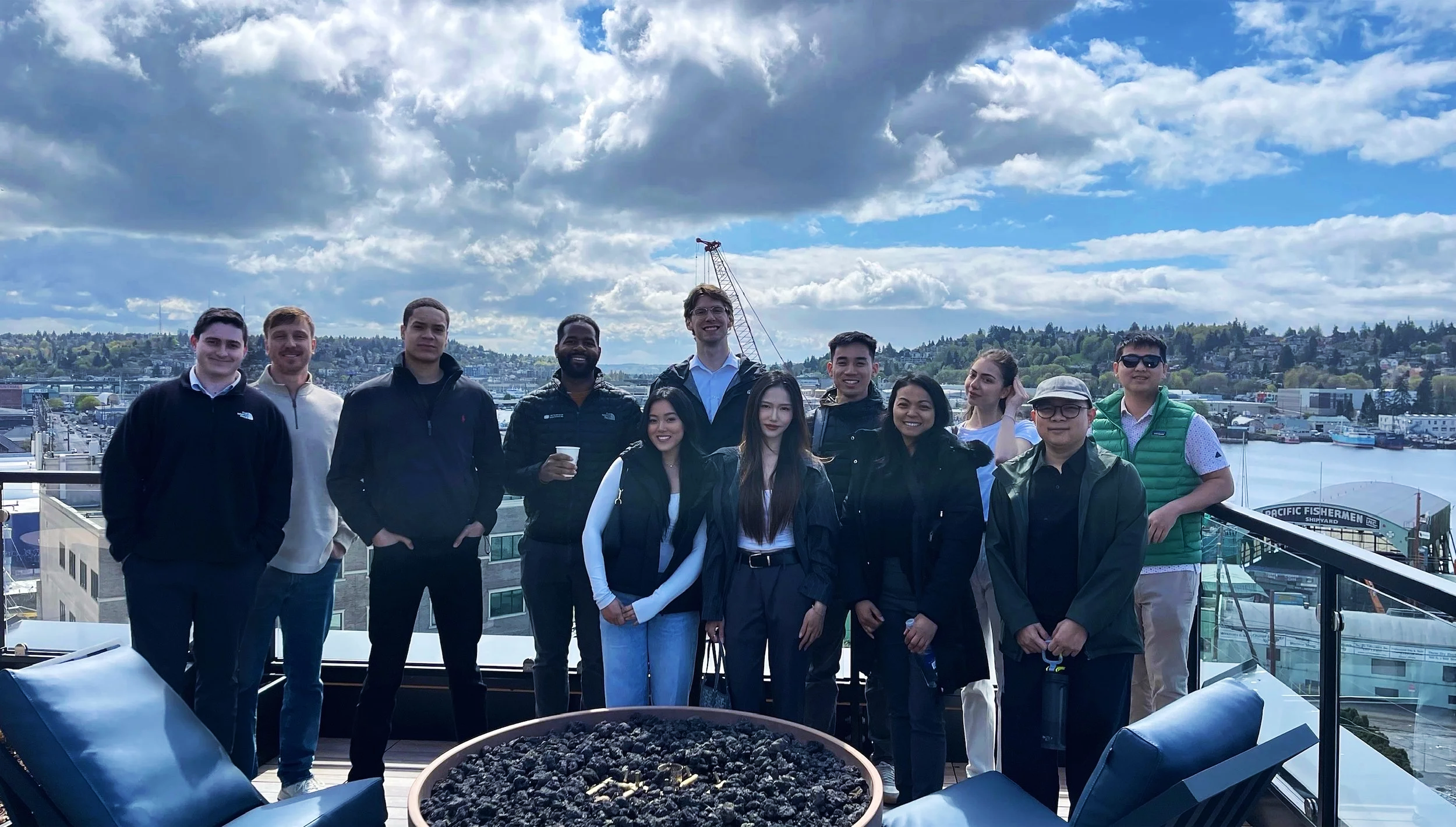 Group of diverse people standing on a rooftop terrace with cityscape and waterfront in the background, under partly cloudy sky.