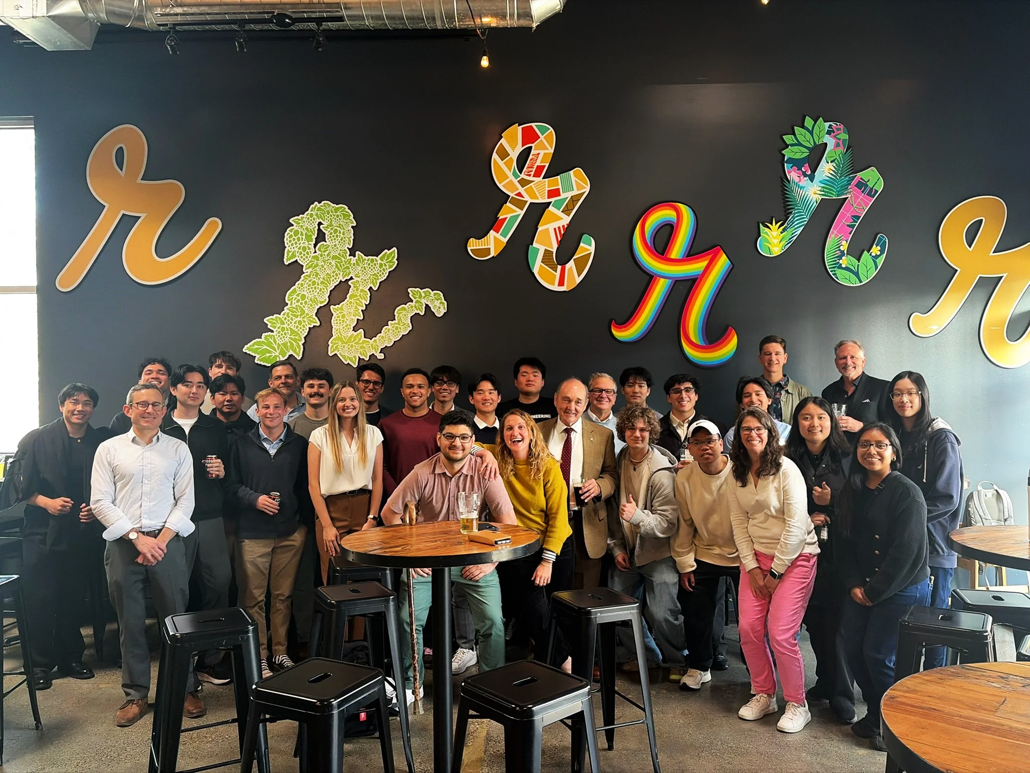 Group of diverse people gathered together in a restaurant or bar, smiling and posing for a photo. There are decorative letters on the wall behind them spelling 'H ...' with colorful and floral designs, and a dark wall with hanging lights.