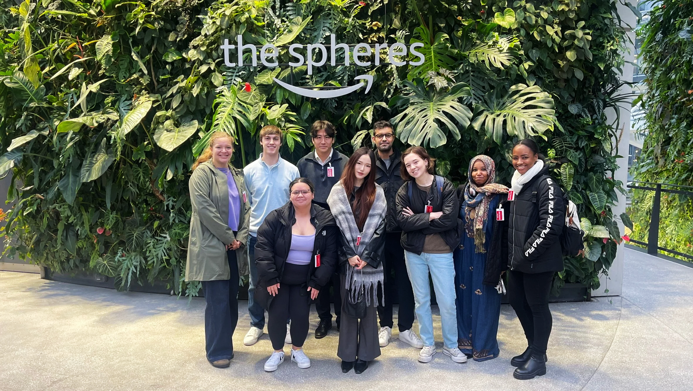 A diverse group of ten people standing in front of a large green wall with the words 'the spheres' and an arrow logo. The group includes men and women with different ethnicities, smiling and looking at the camera, in an indoor space.