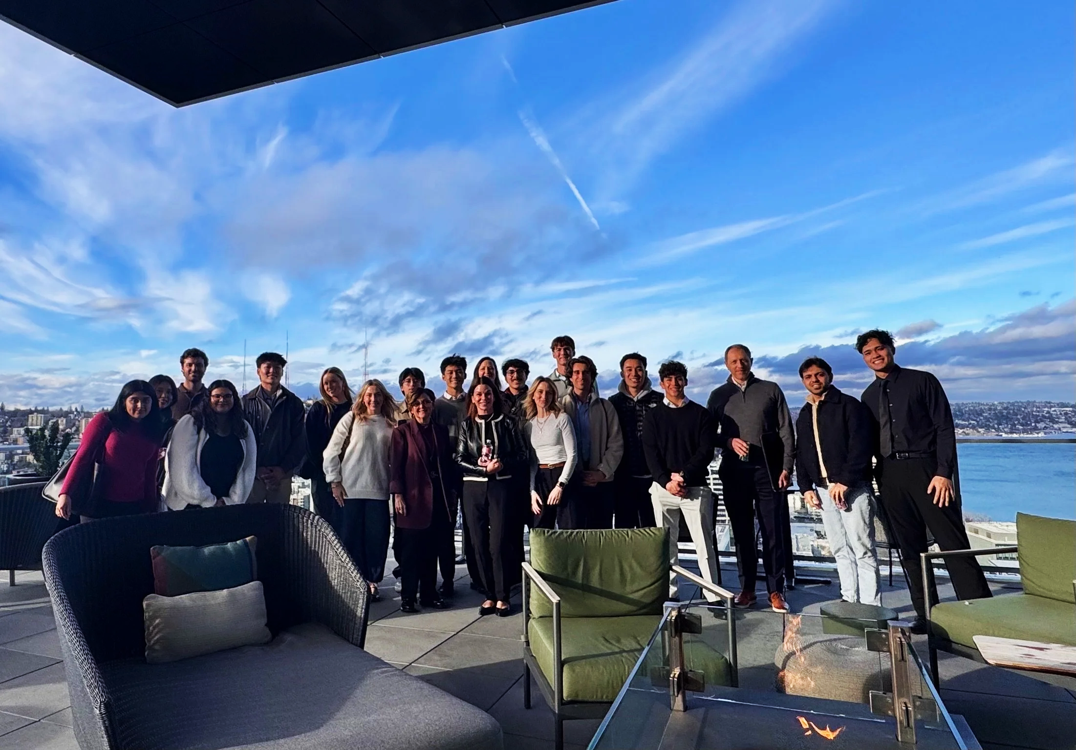 A group of people standing on a rooftop terrace with a view of a body of water and cityscape in the background, during daytime with partly cloudy sky.