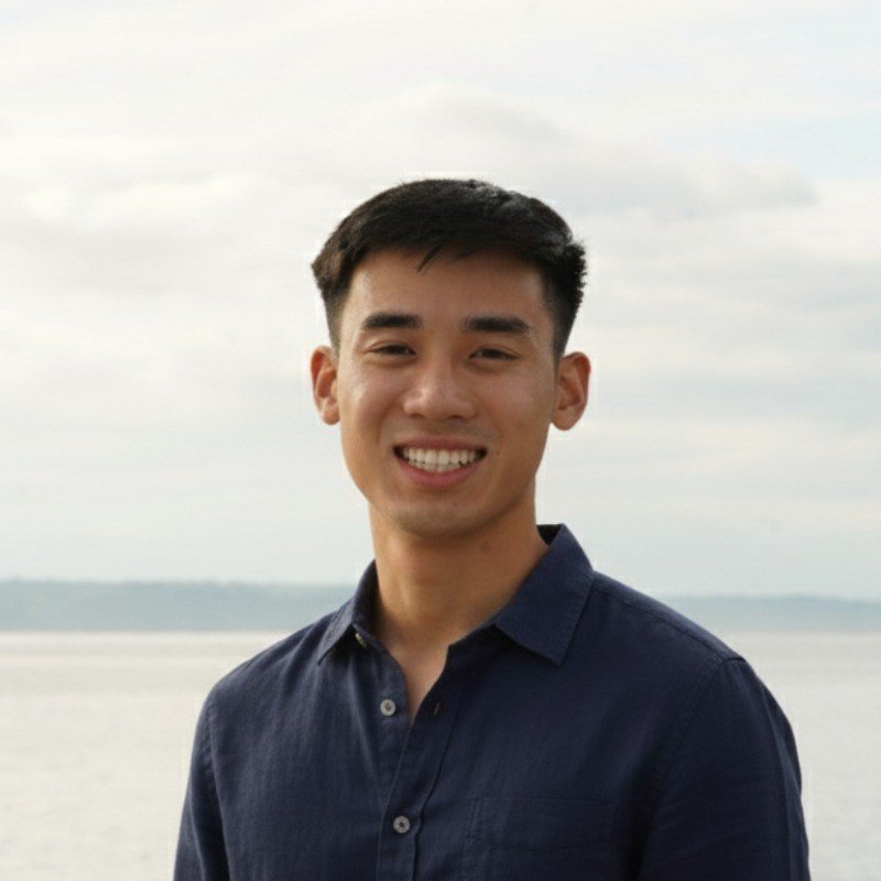 Young man smiling outdoors near a body of water with a cloudy sky.