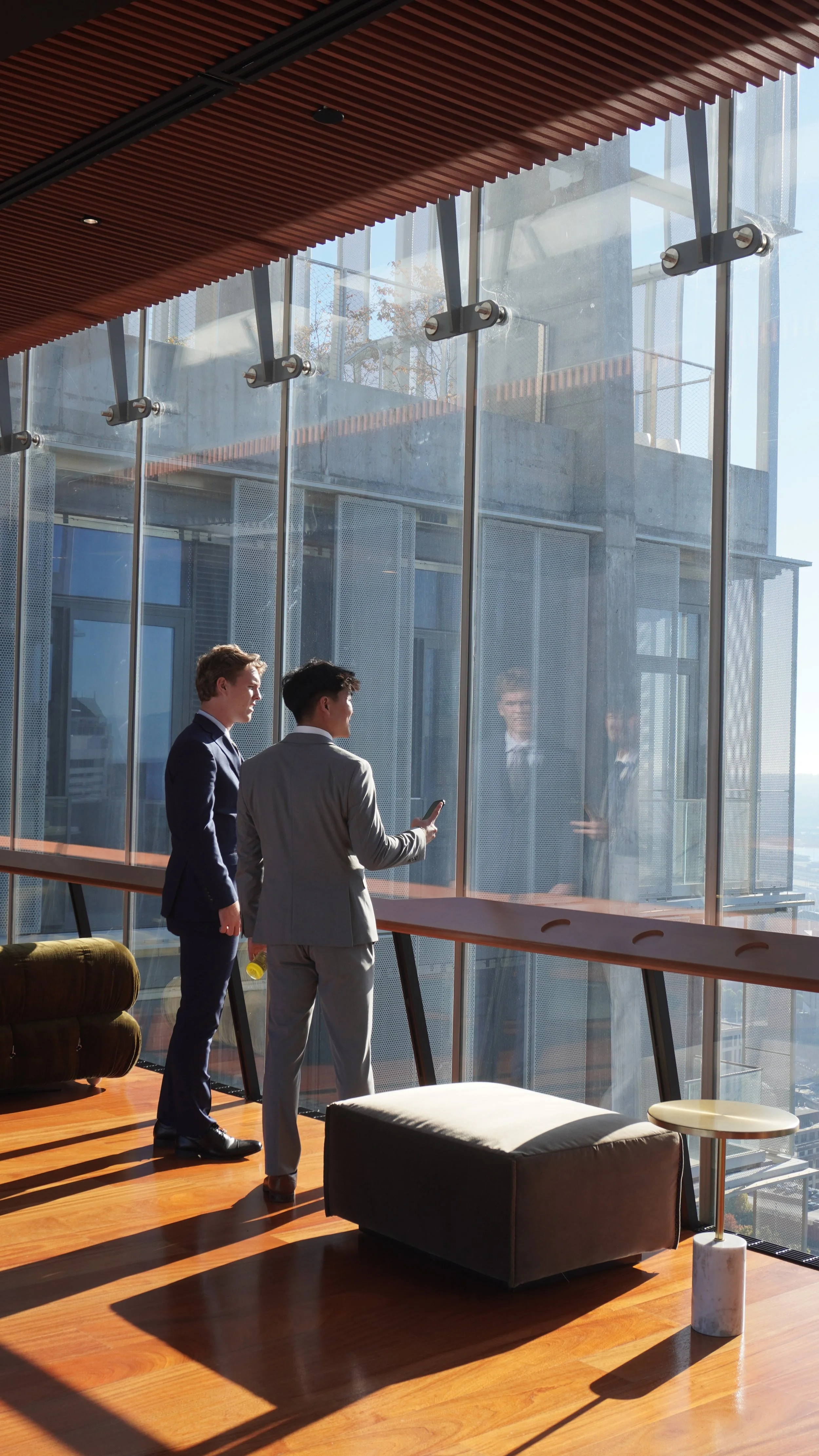 Two businessmen in suits standing in a modern office with large glass windows, one showing something on his phone to the other, and their reflections visible in the window.