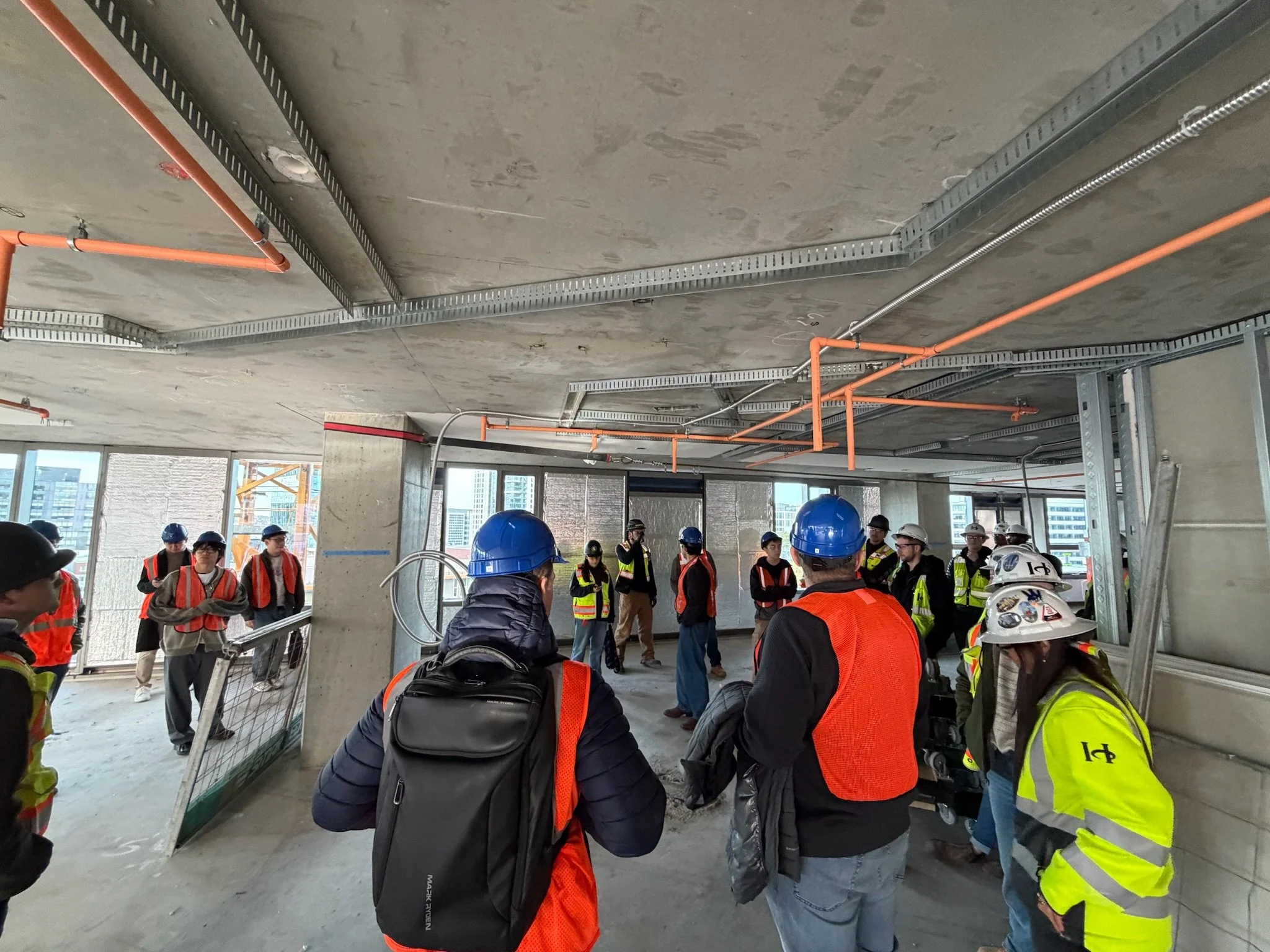 A group of construction workers wearing safety helmets and vests gathering inside a building under construction, with exposed pipes and metal framework on the ceiling and large windows in the background.
