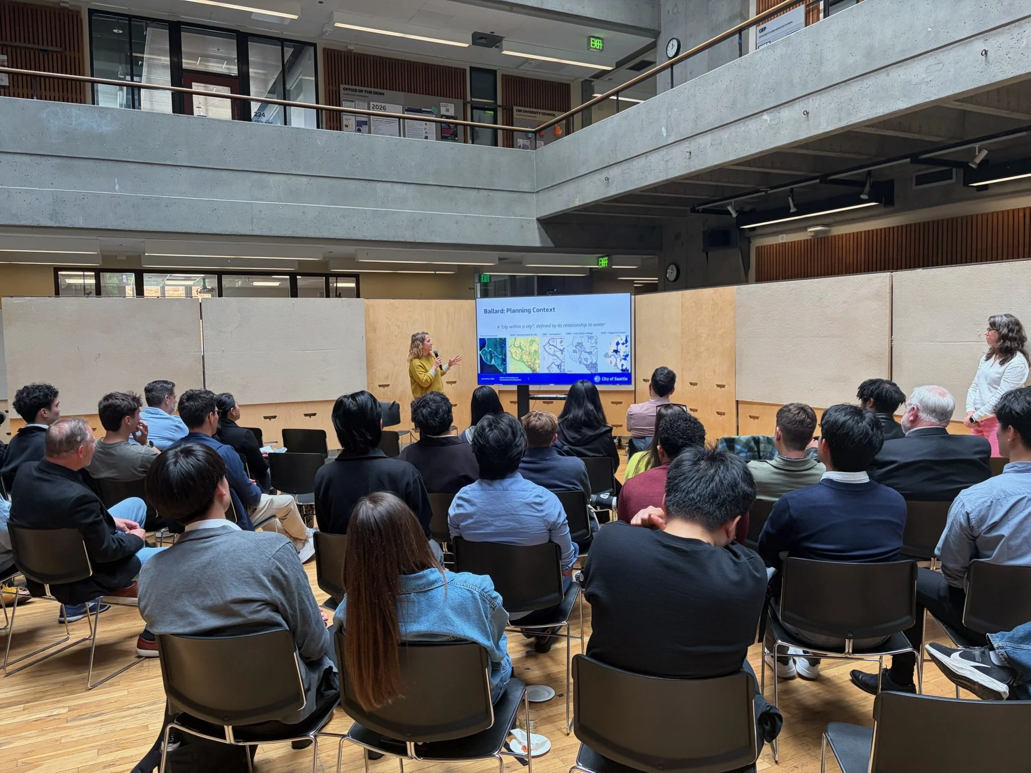 A woman giving a presentation to an audience in a modern, multi-level indoor setting. The presentation slide on the screen mentions 'Ballard: Planning Context' and is attributed to the City of Seattle.