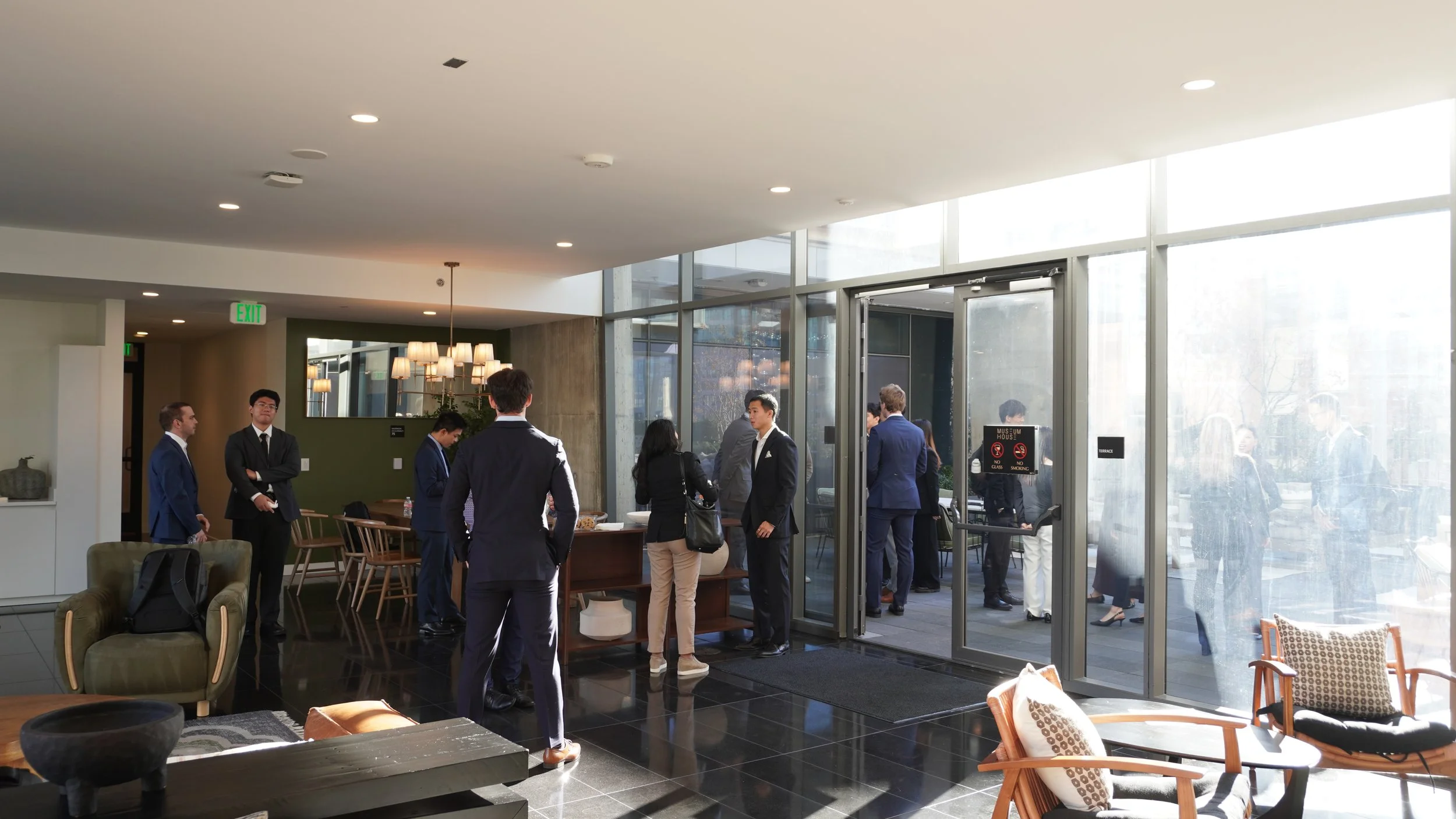 Business people in suits gathered inside a modern hotel lobby with large glass doors, relaxing chairs, and a stylish interior design.