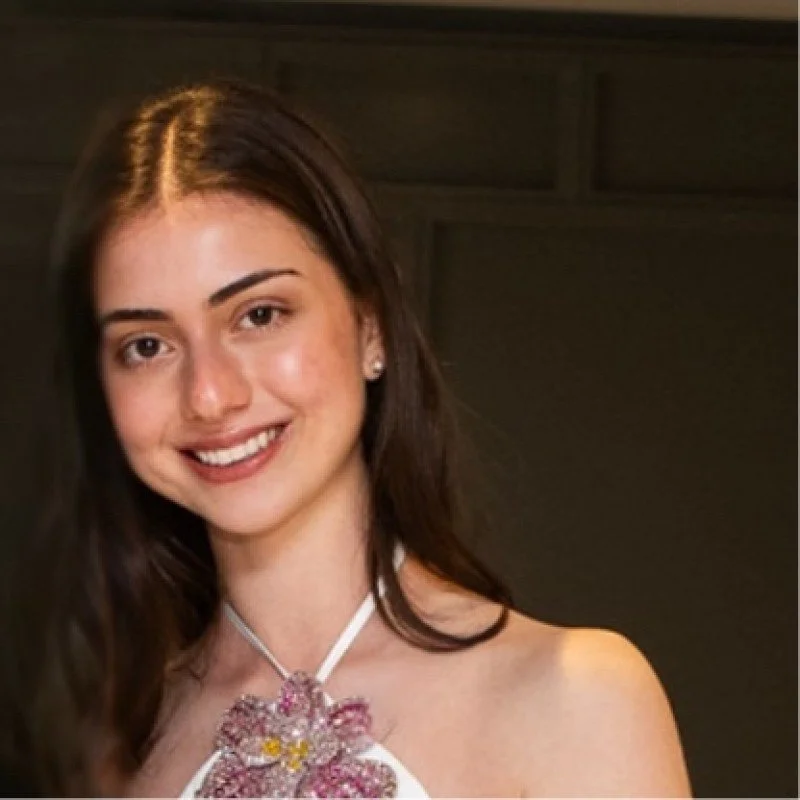 Young woman with long dark hair, wearing a floral necklace and a sleeveless top, smiling at the camera.