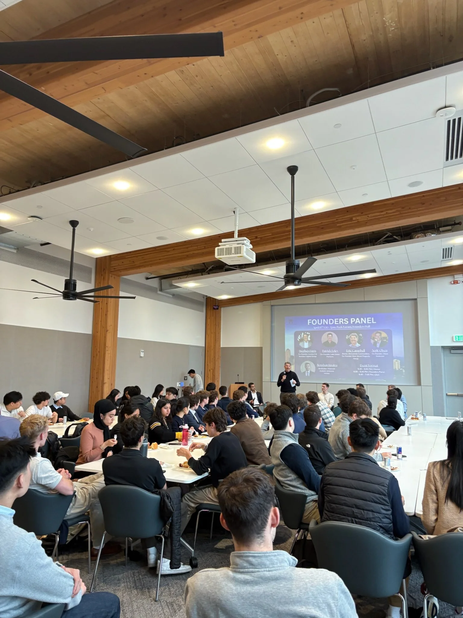 A large group of people attending a panel discussion event in a conference room with a wooden ceiling and modern decor. The panel is displayed on a large screen showing speakers' photos and names.