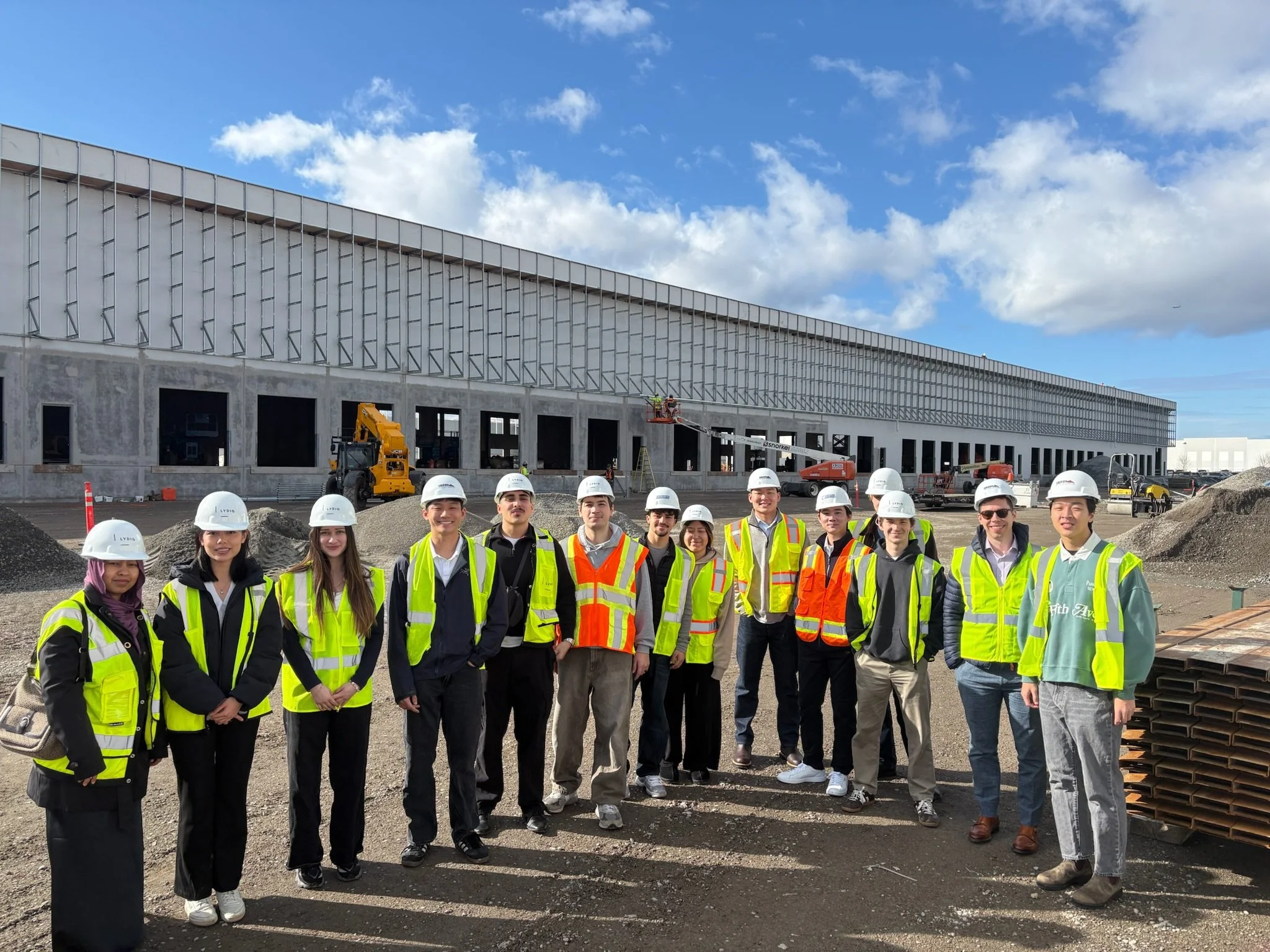 Group of construction workers and students wearing safety helmets and reflective vests on a construction site with building framework and blue sky in the background.