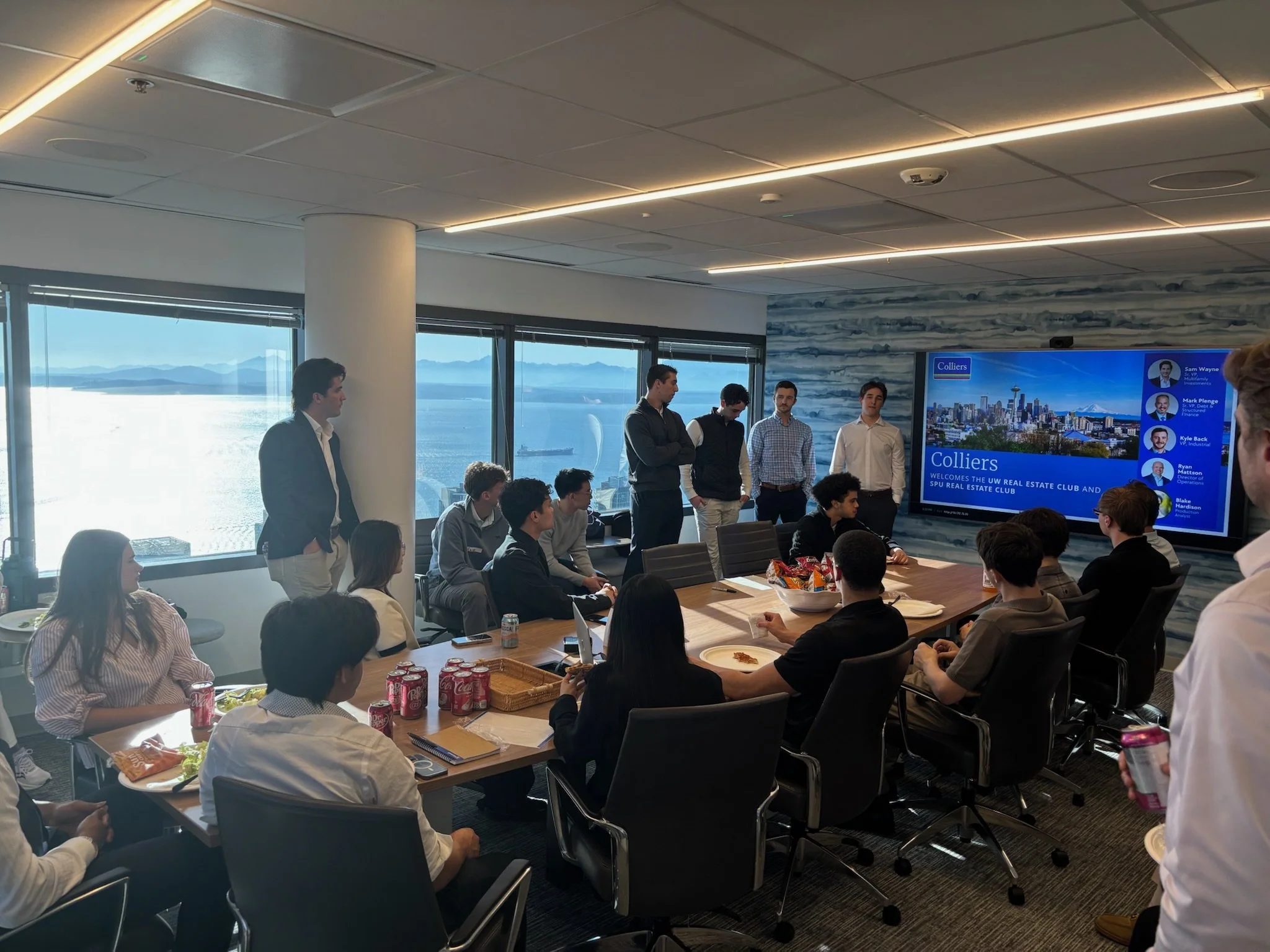 A group of people in business casual attire attending a presentation or meeting in a conference room with large windows showing a city and water view. Some are seated, others are standing near a large screen displaying a presentation about a real est