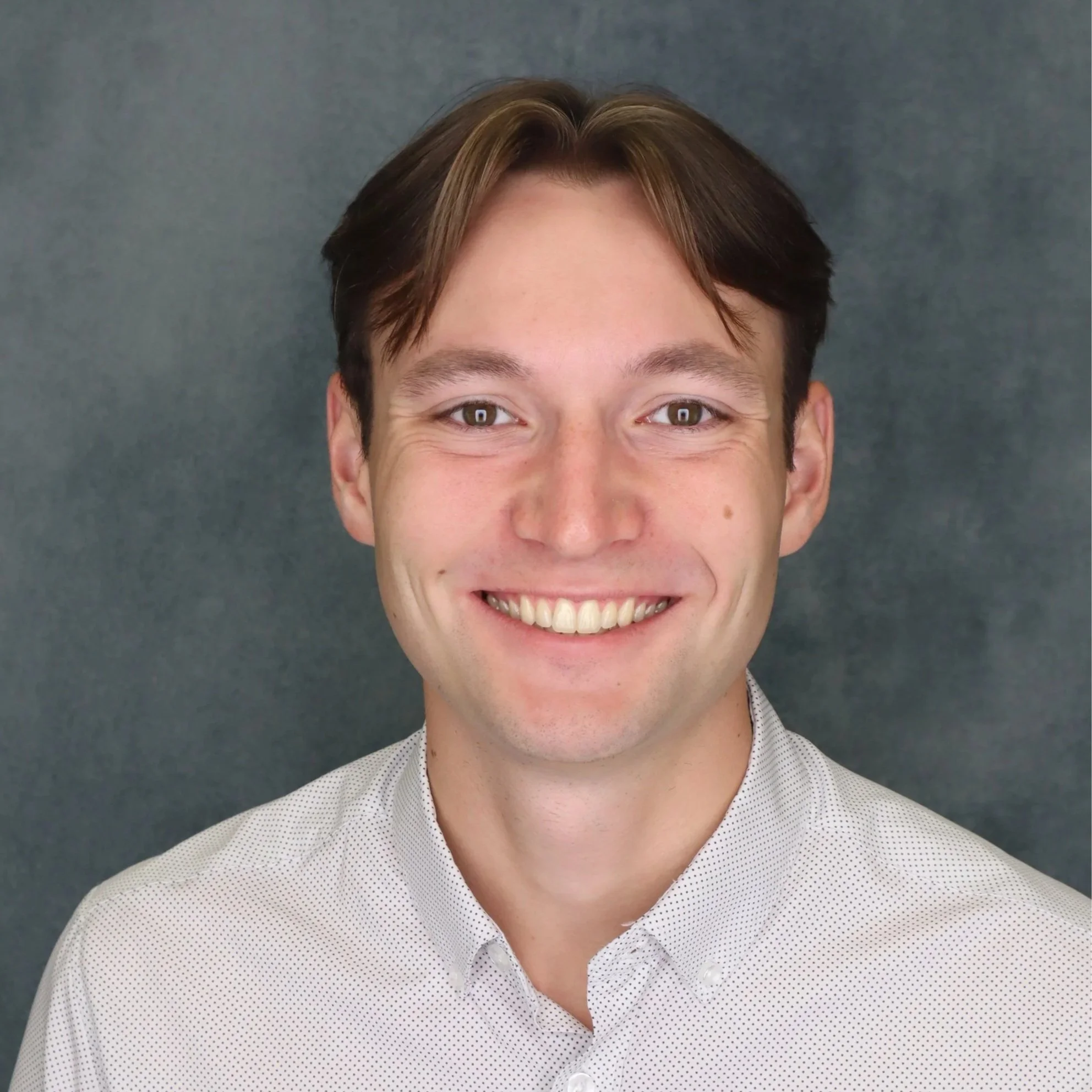 Close-up of a smiling man with short brown hair, green eyes, wearing a light-colored polka-dot shirt, against a dark gray background.