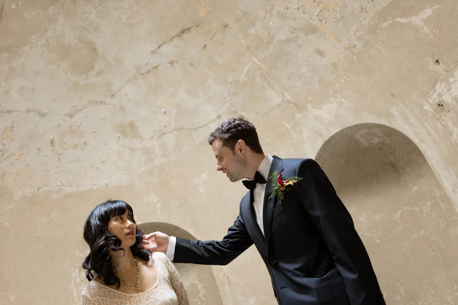 A groom in a black tuxedo with a boutonniere touches the face of a bride in a lace dress during a wedding.