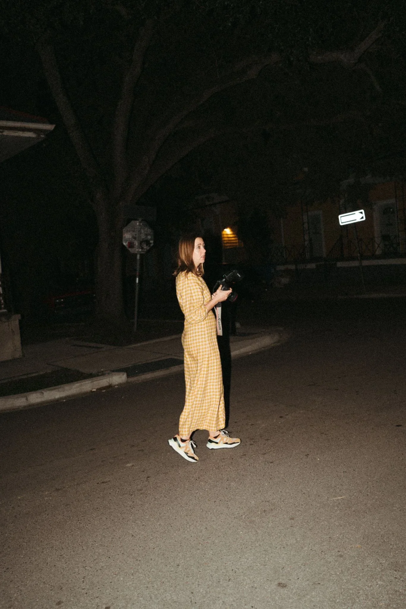 A woman wearing a yellow checkered jumpsuit and sneakers walking on a dark street at night while holding a camera.