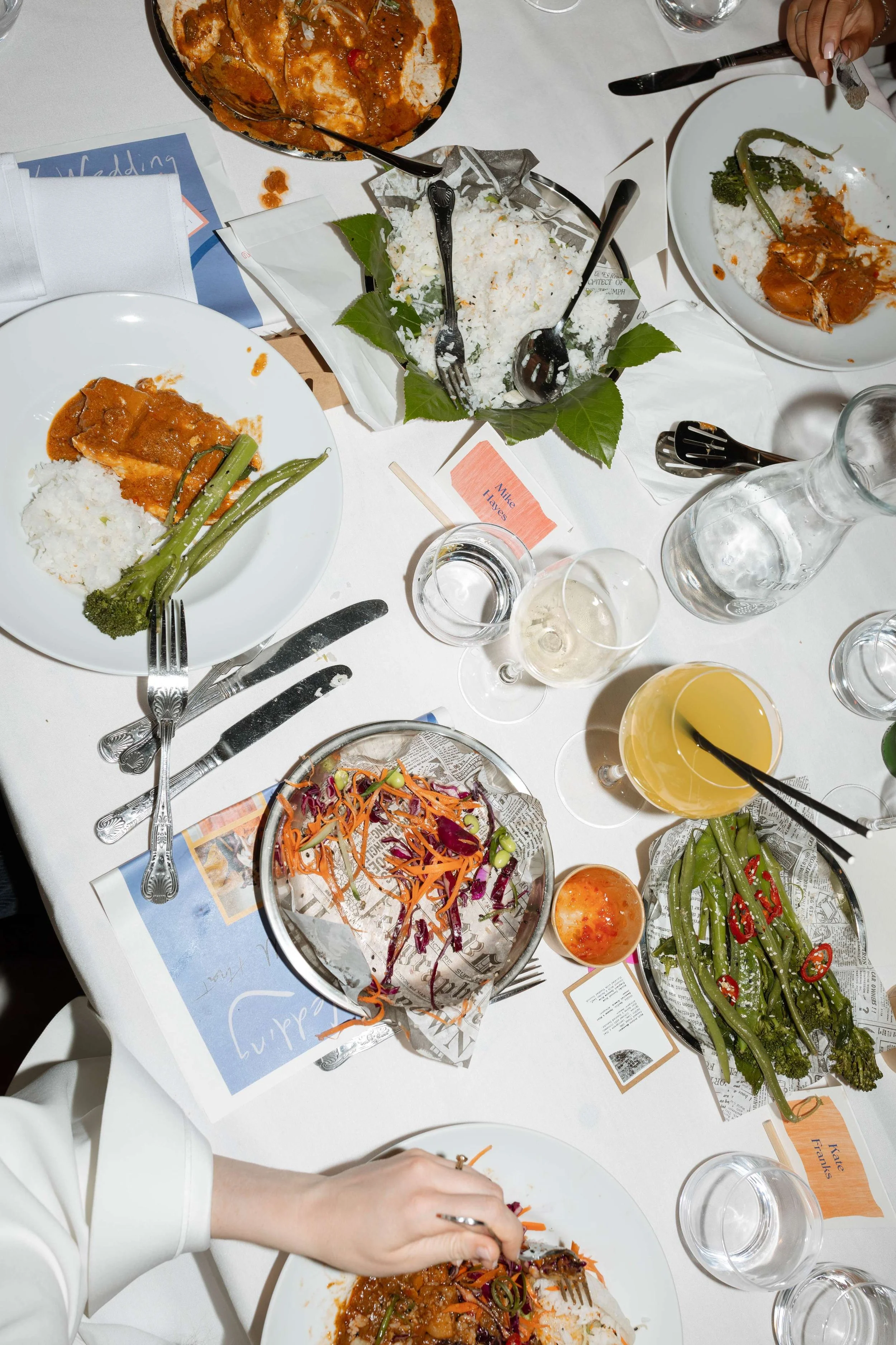 A table with partially eaten plates of food, including rice and curry, green beans, a salad with shredded vegetables, and drinks such as white wine, champagne, and orange juice. There are utensils, glasses, and decorative leaves on the table.