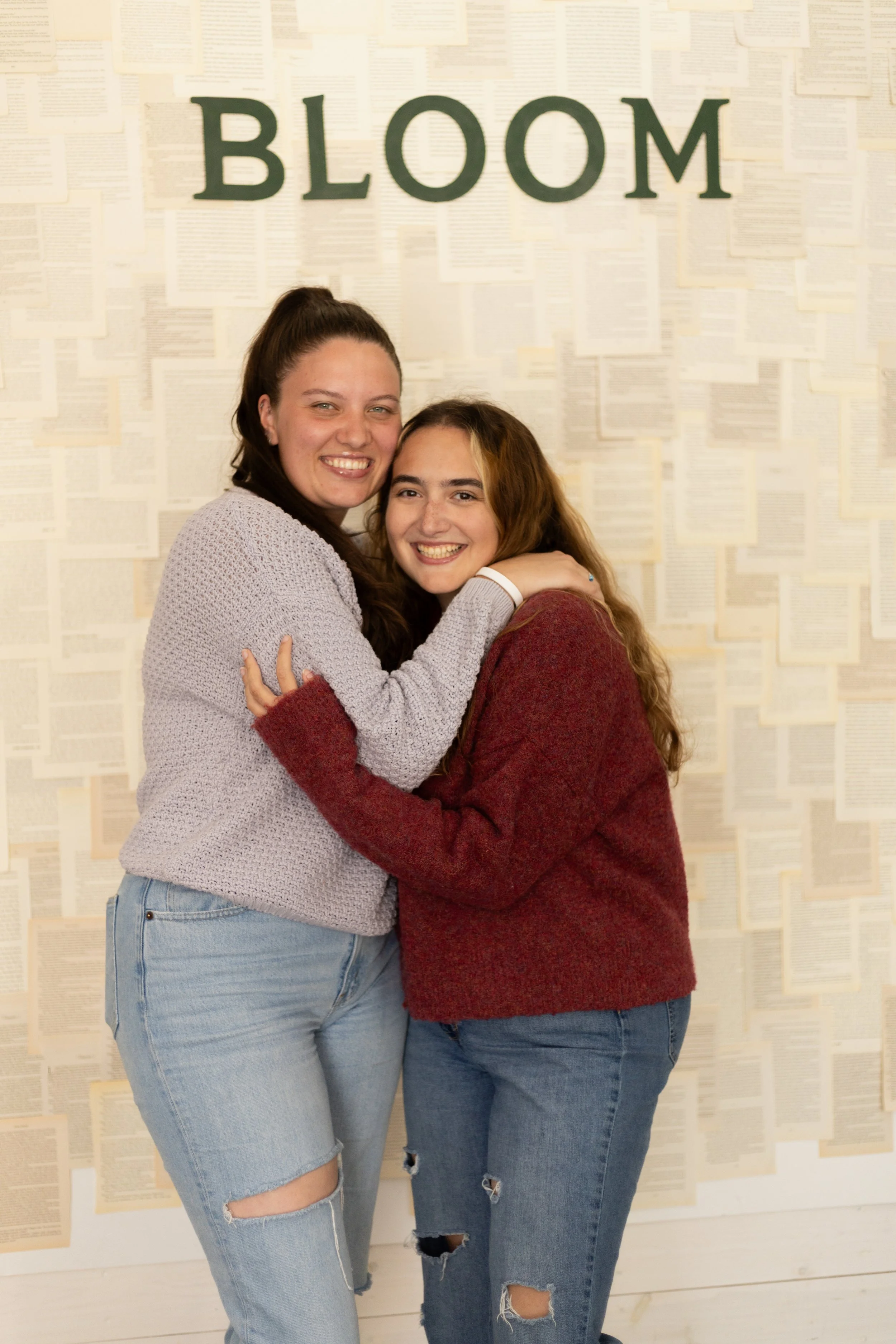Two young women hugging and smiling in front of a wall with pages of text and a sign that says 'BLOOM'.