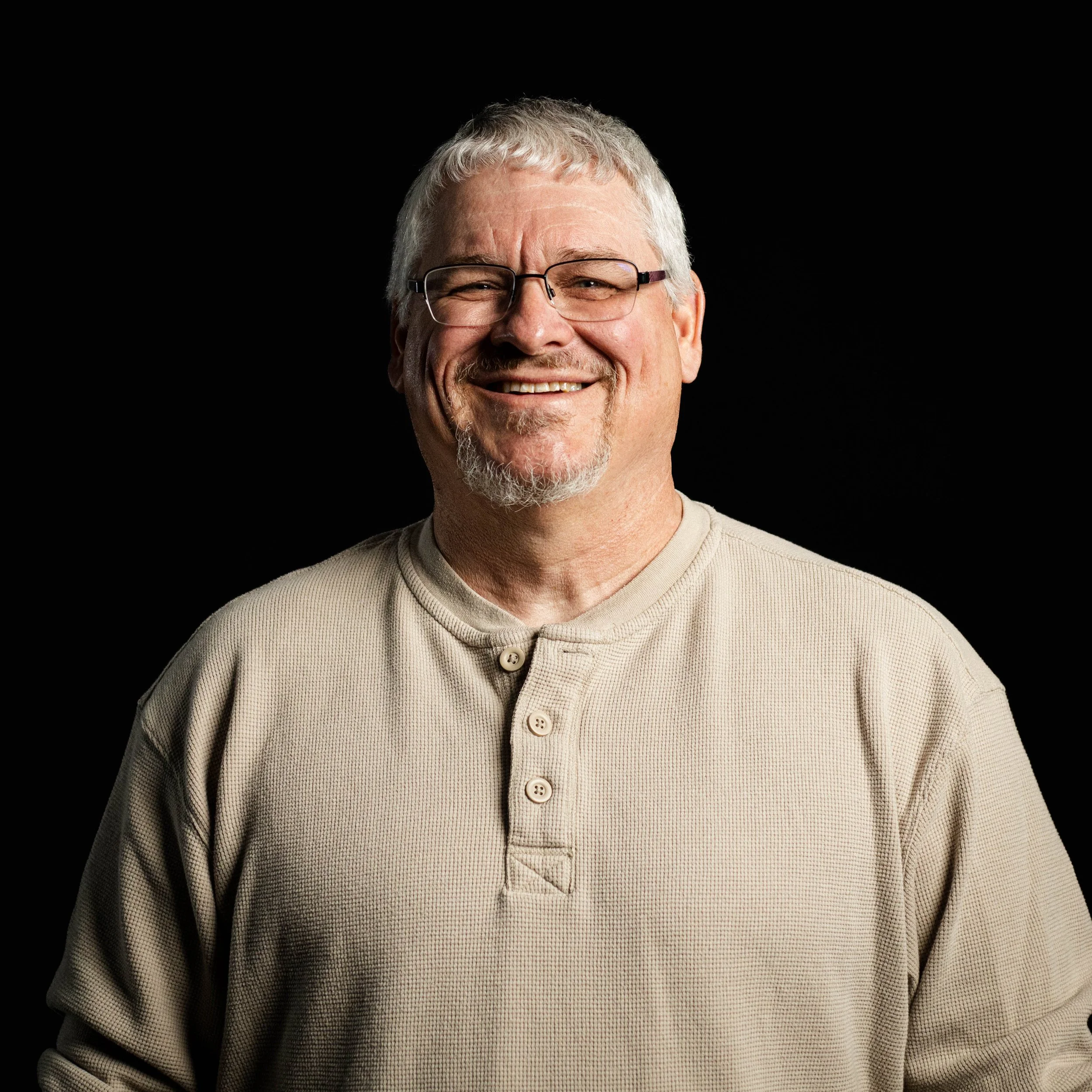 A smiling middle-aged man with gray hair, glasses, and a beard, wearing a beige long-sleeve shirt, standing against a black background.