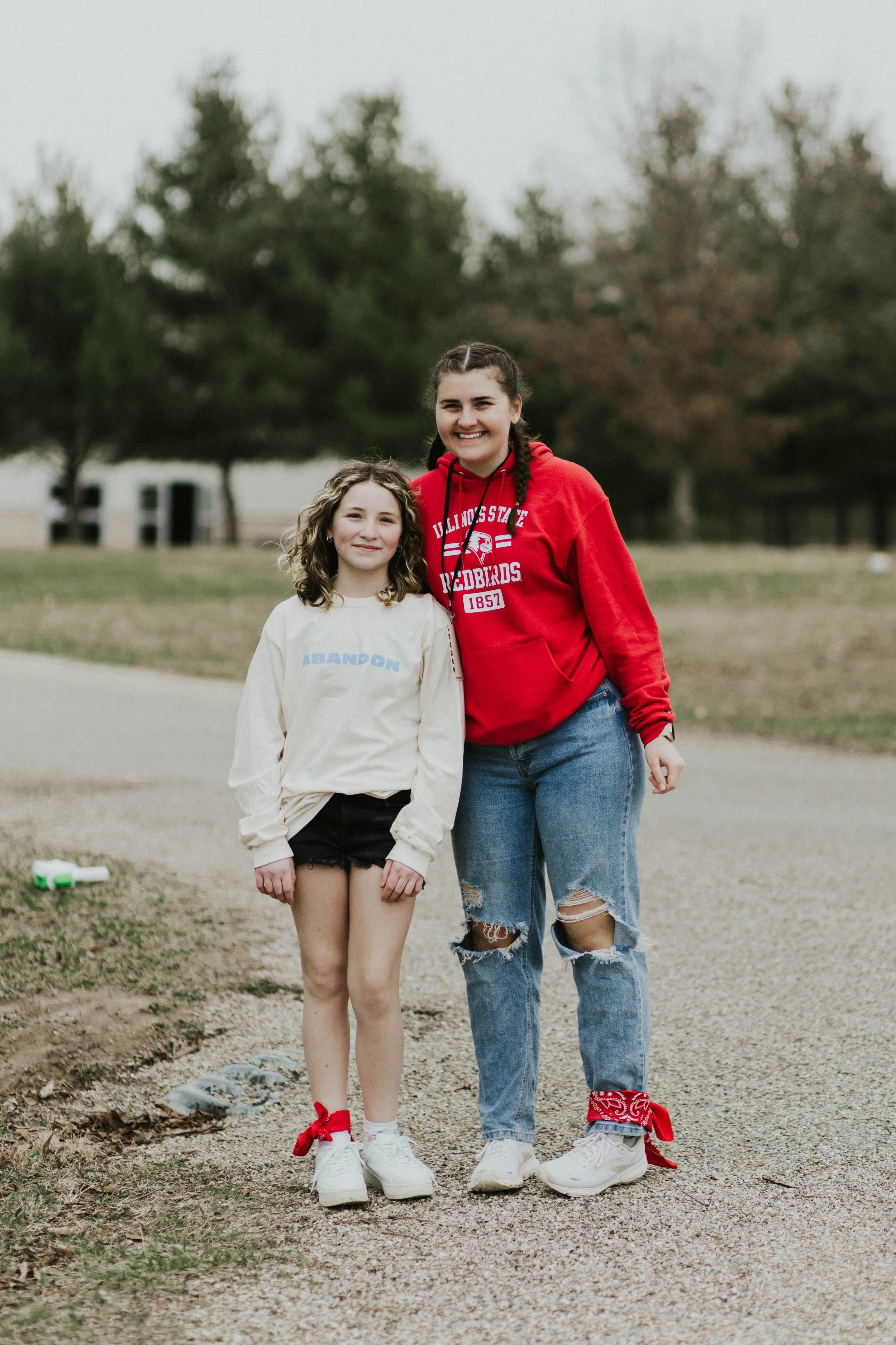 Two girls standing outdoors on a dirt path, smiling. One girl is wearing a red hoodie and ripped jeans, the other girl is wearing a cream-colored sweatshirt and black shorts. Both are wearing white sneakers with red bandanas tied around their ankles.
