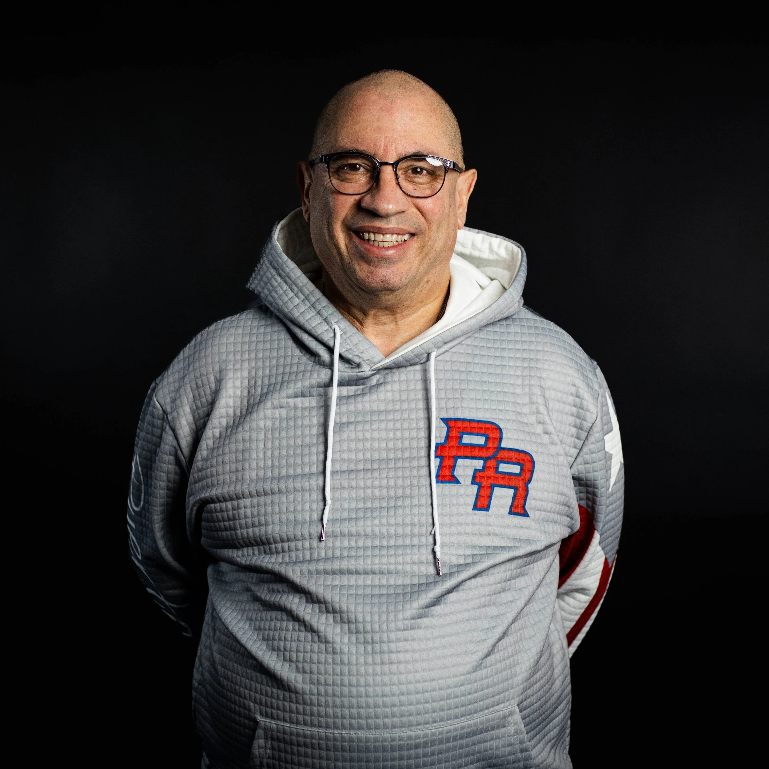 An elderly man with glasses, wearing a red cardigan over a checkered shirt, standing against a black background.