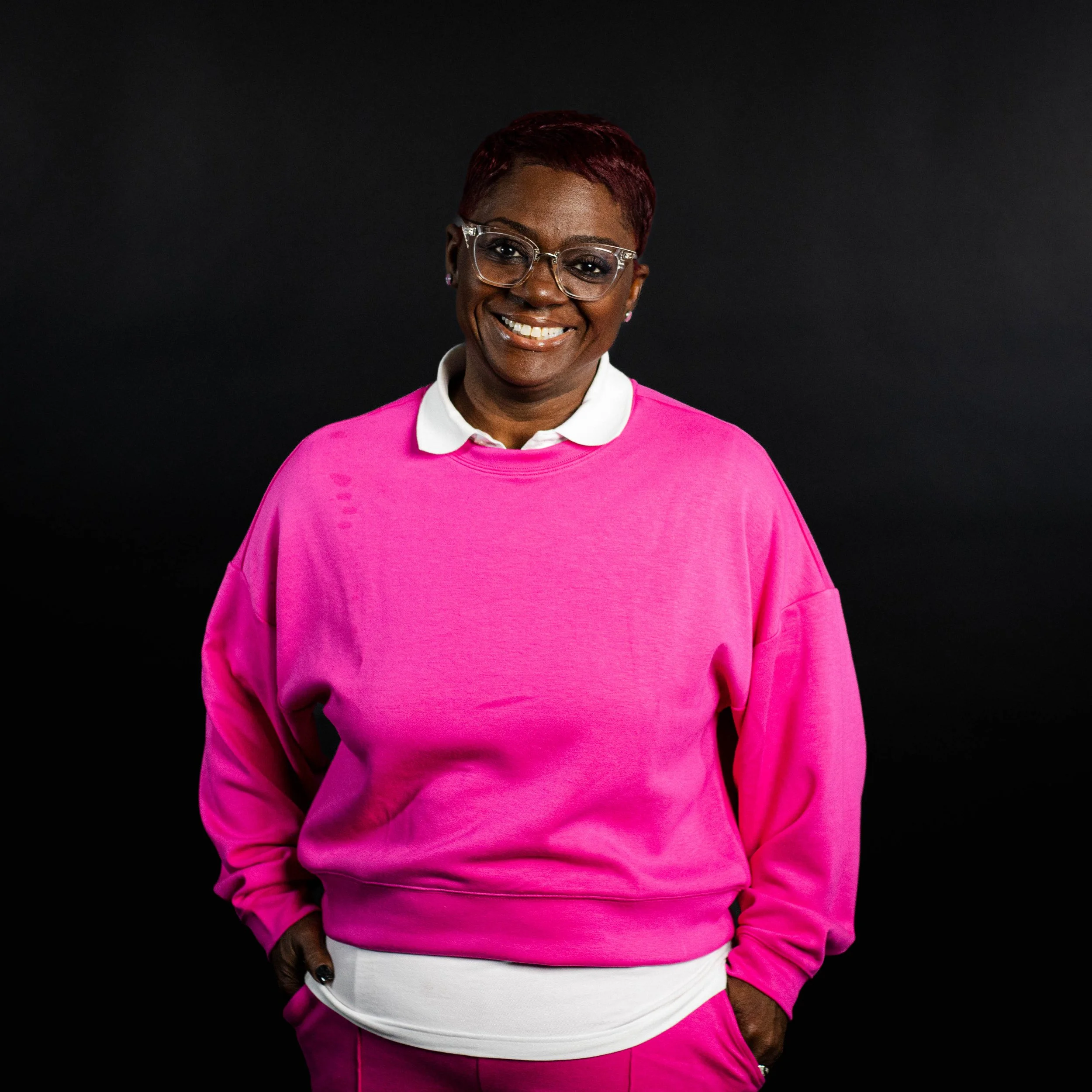 A woman with dark skin, short red hair, and glasses wearing a bright pink sweatshirt over a white collared shirt, smiling against a black background.
