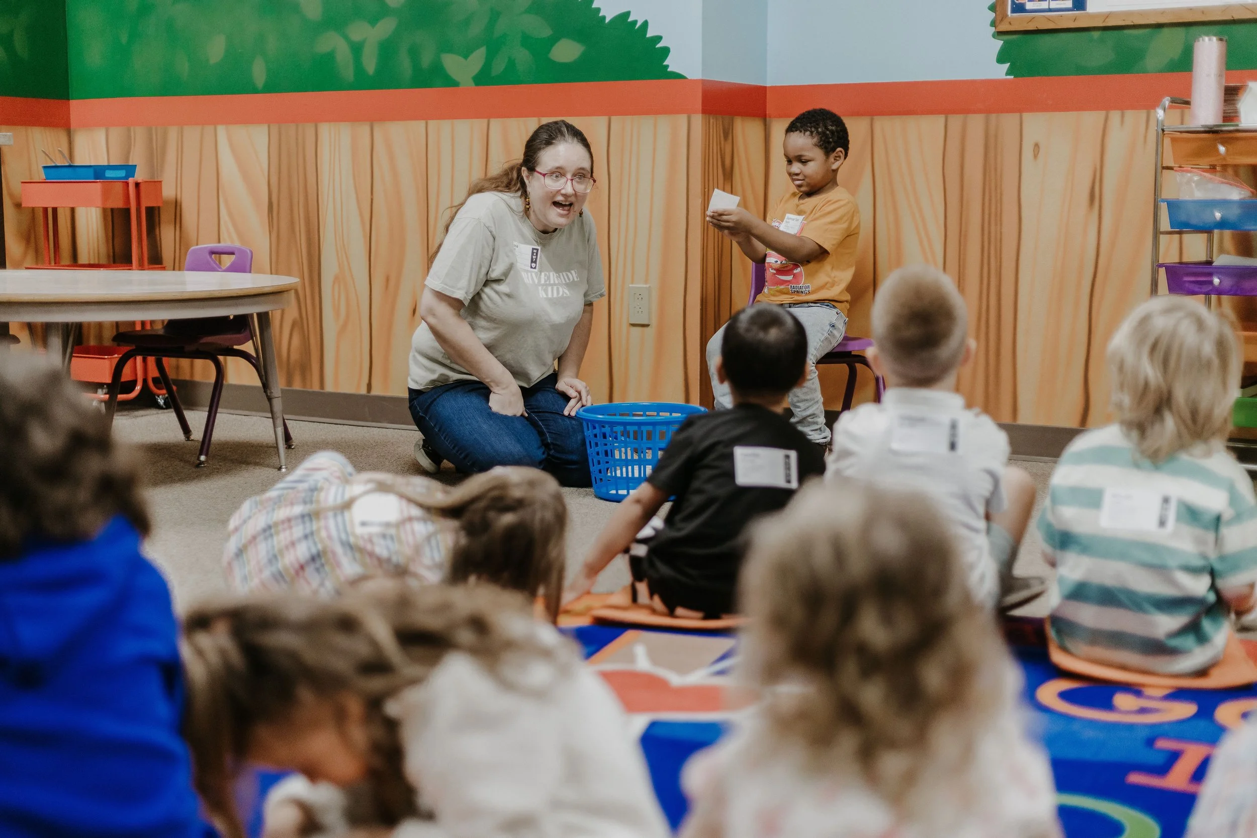 A woman with glasses and long hair is kneeling on the floor, smiling and engaging with children in a colorful indoor setting. A young boy with curly hair is sitting on a purple chair, holding a card or paper. Several children are seated on the floor, facing the woman and boy, some with badges or tags. The room has bright, playful wall decorations with trees and a sorting cart with colored bins.