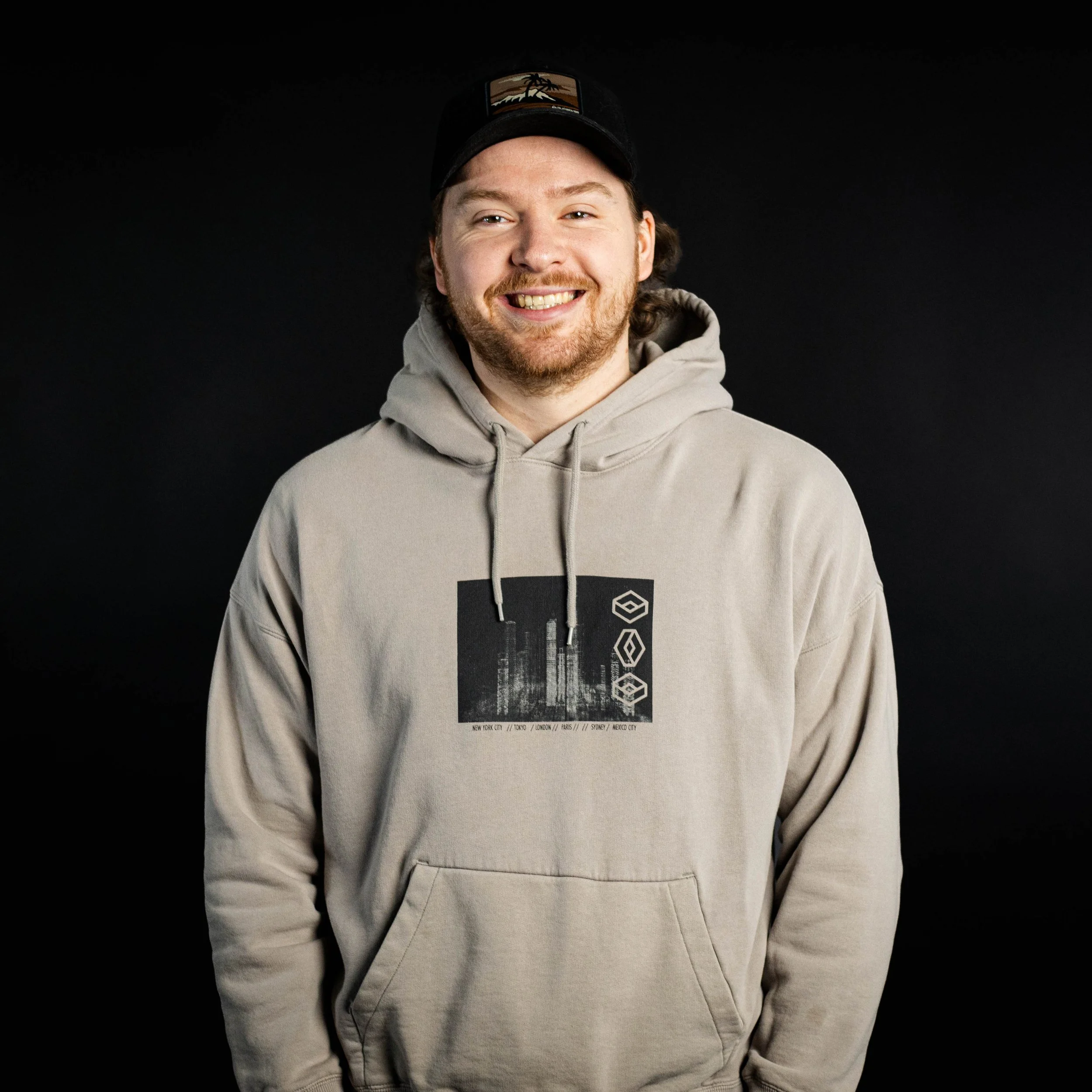 A young man with light skin, curly brown hair, and a beard, smiling and wearing a beige hoodie with a graphic design on the front, and a black baseball cap against a black background.