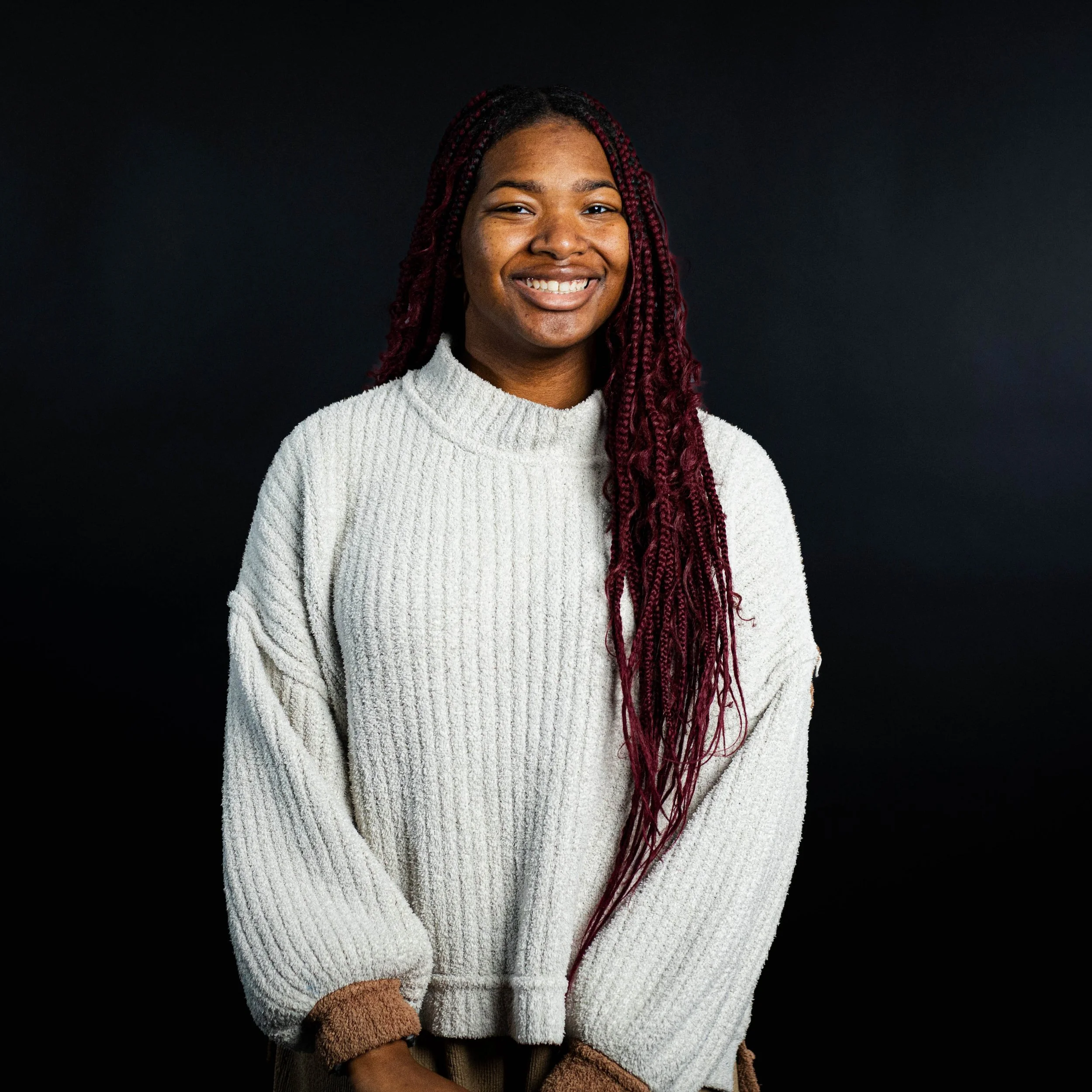 A young woman with long, reddish braids smiling in front of a black background, wearing a white sweater.
