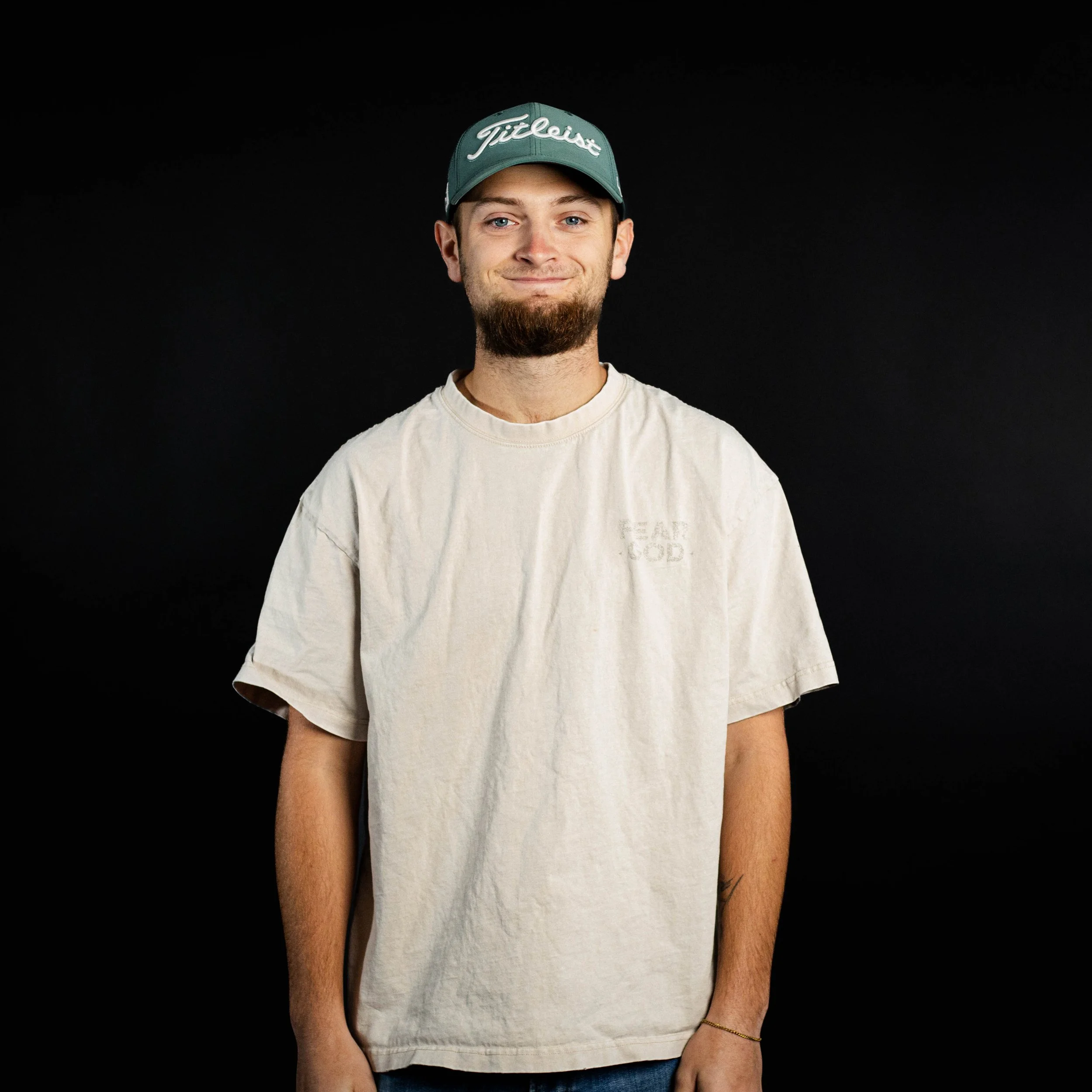 A young man with a beard and mustache wearing a beige T-shirt and a green golf cap, smiling at the camera against a black background.